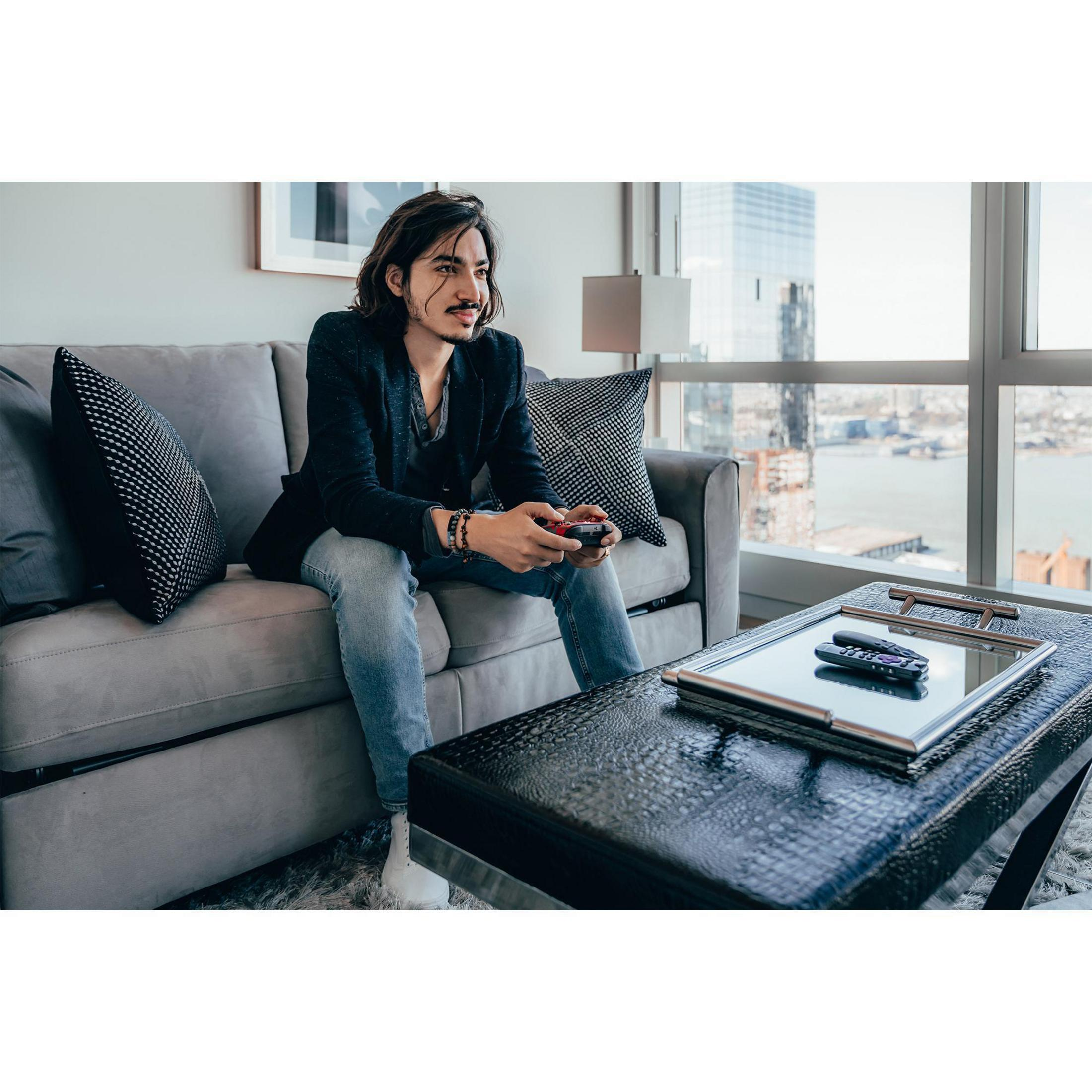 Mann sitzt auf einem Sofa und spielt ein Videospiel. Er hält einen Controller. Hintergrund: Fenster, Tisch.