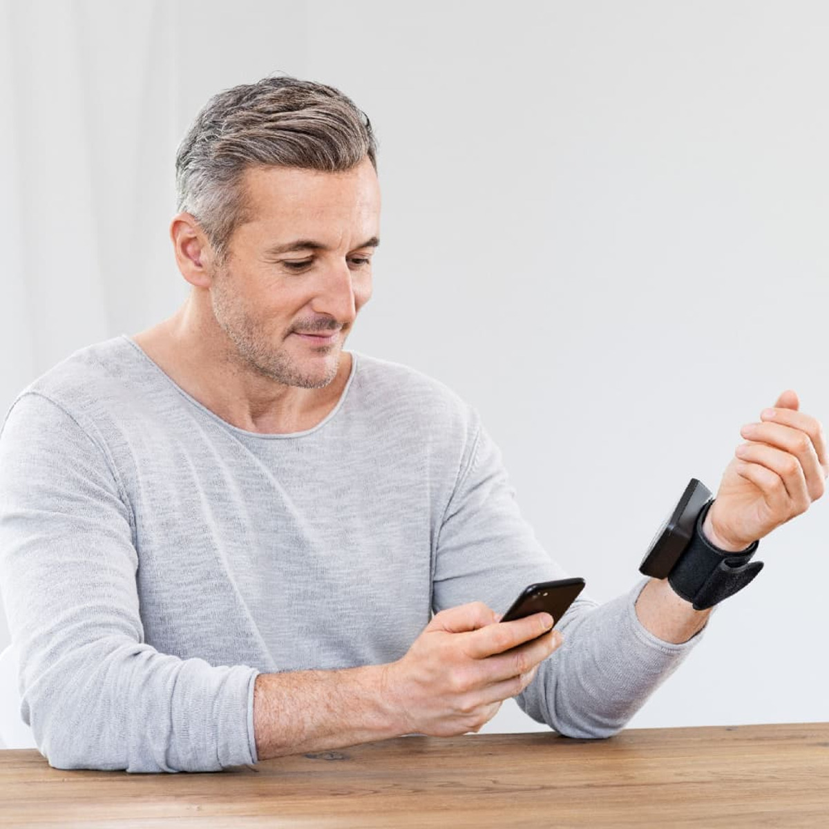 Mann hält Telefon, trägt ein schwarzes Armband. Holztisch. Weißer Hintergrund.
