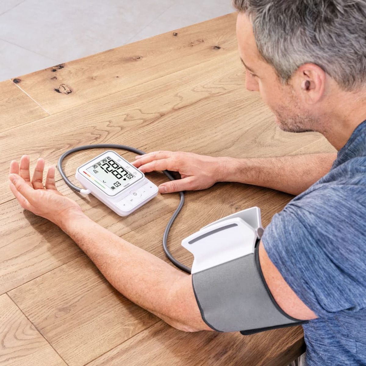 Ein Mann misst seinen Blutdruck mit einem digitalen Blutdruckmessgerät auf einem Holztisch.