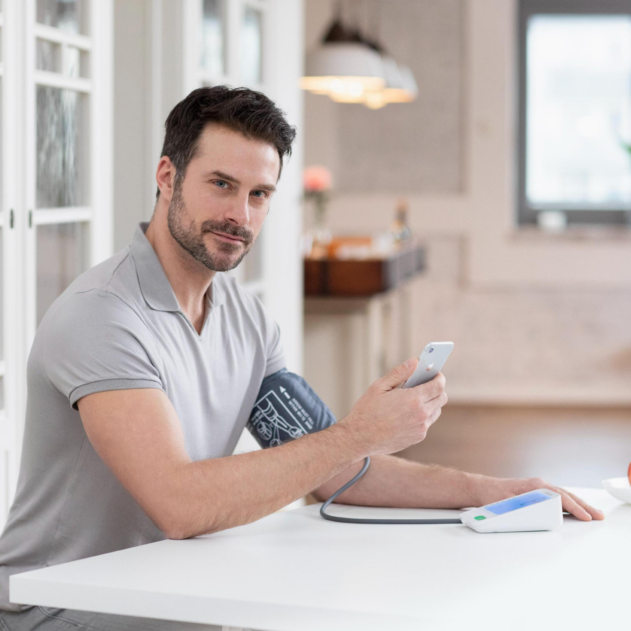 Mężczyzna z monitorem ciśnienia na ramieniu, trzymający telefon, biały stół, dom.