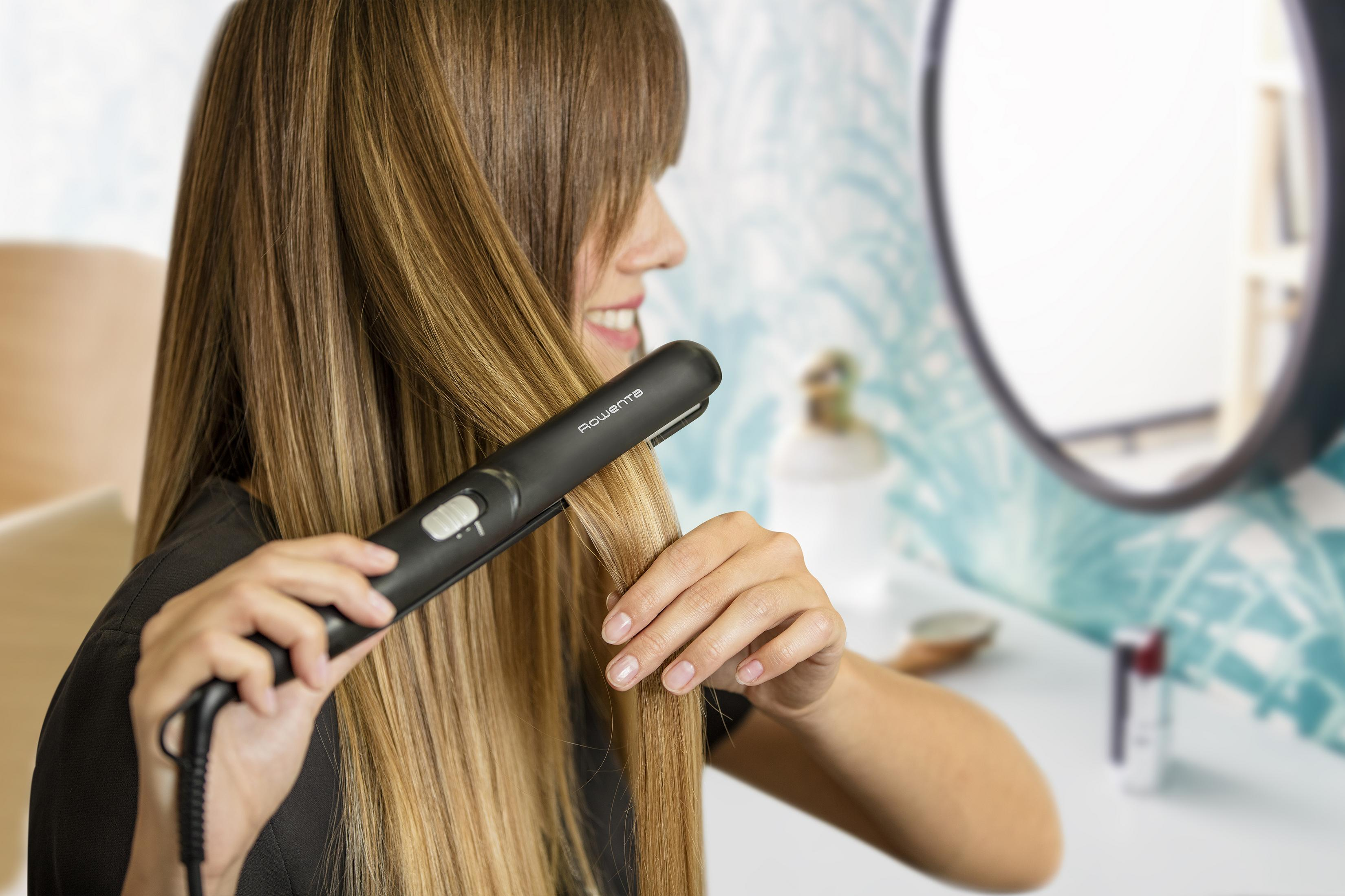 Kobieta używa czarnej prostownicy do długich, jasnobrązowych włosów przed lustrem.