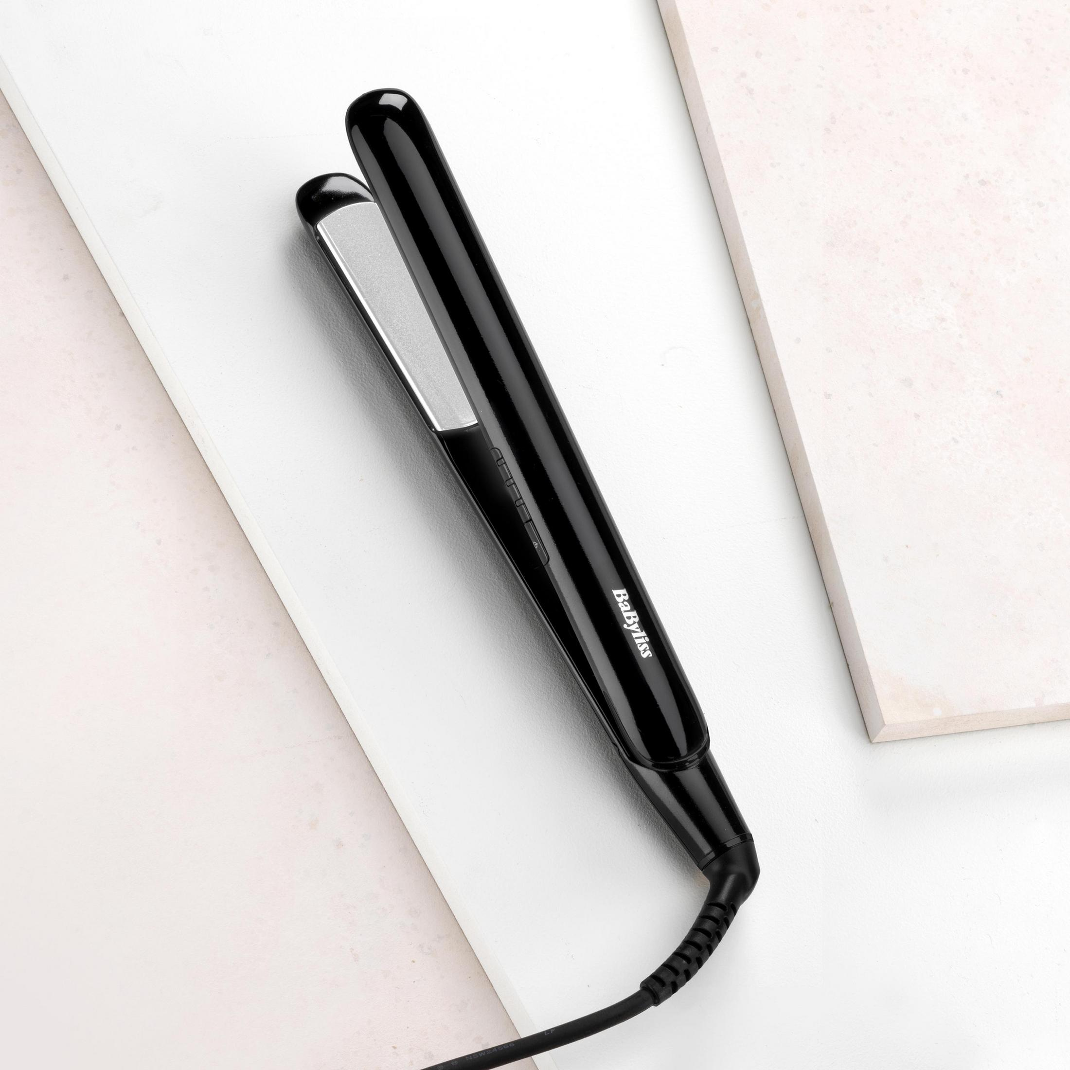 Czarna prostownica do włosów ze srebrnymi płytkami, na białej powierzchni o marmurowej fakturze. Czarny przewód.