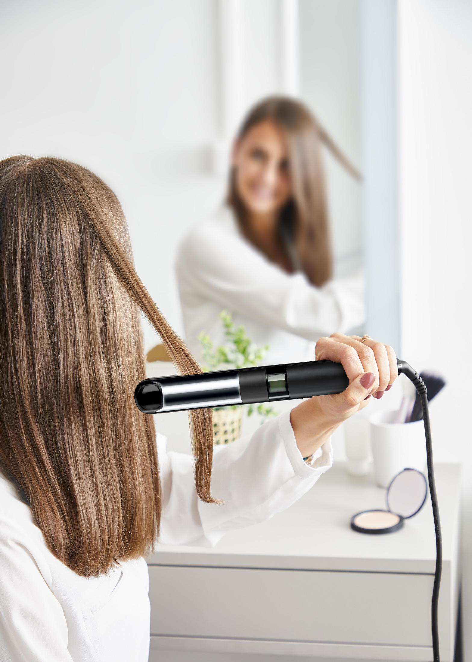 Frau benutzt einen Haarglätter vor einem Spiegel. Eine andere Frau spiegelt sich.