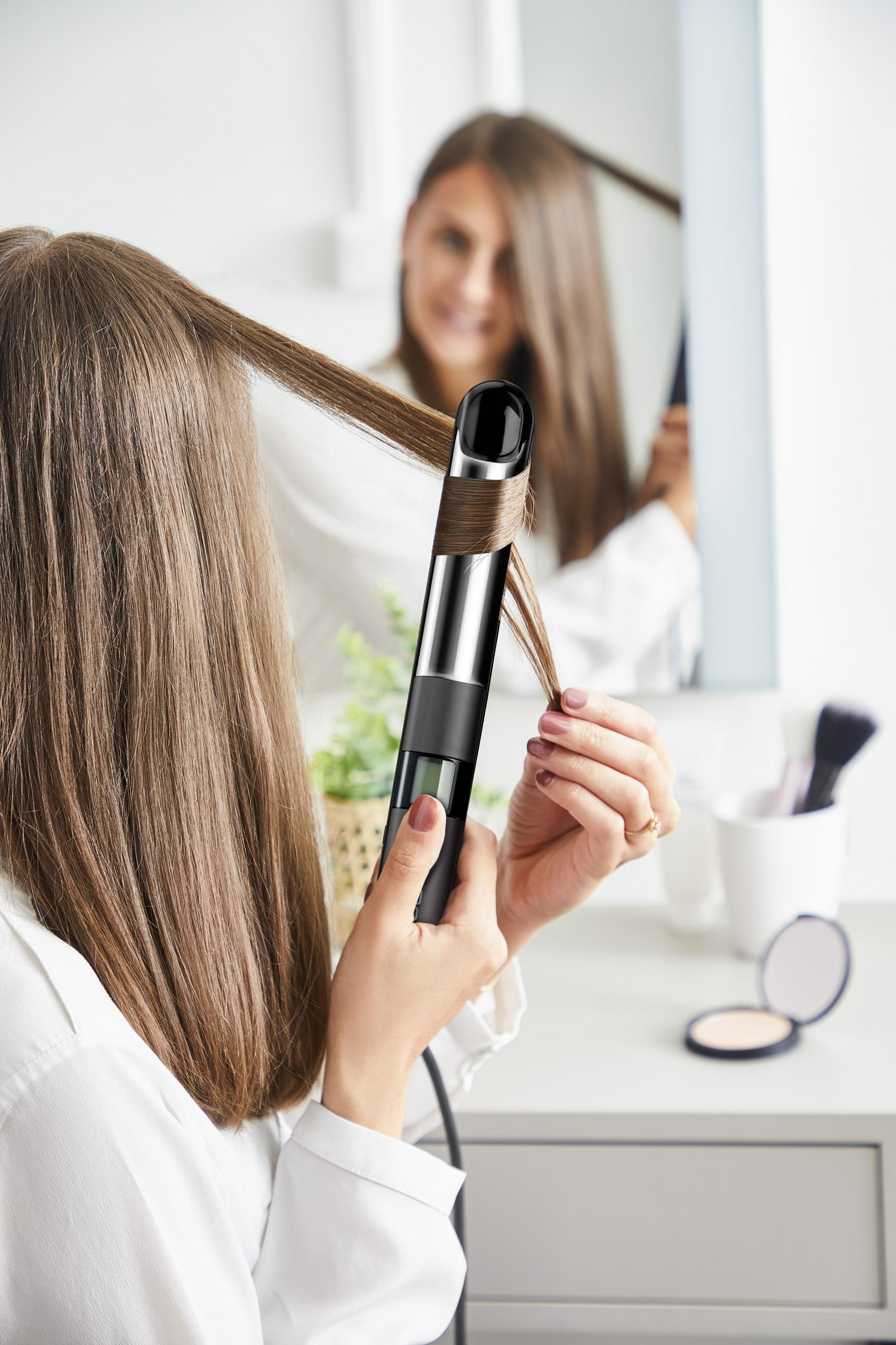 Frau, die sich vor einem Spiegel die Haare mit einem Styling-Tool lockt. Make-up und Produkte stehen auf dem Tisch.