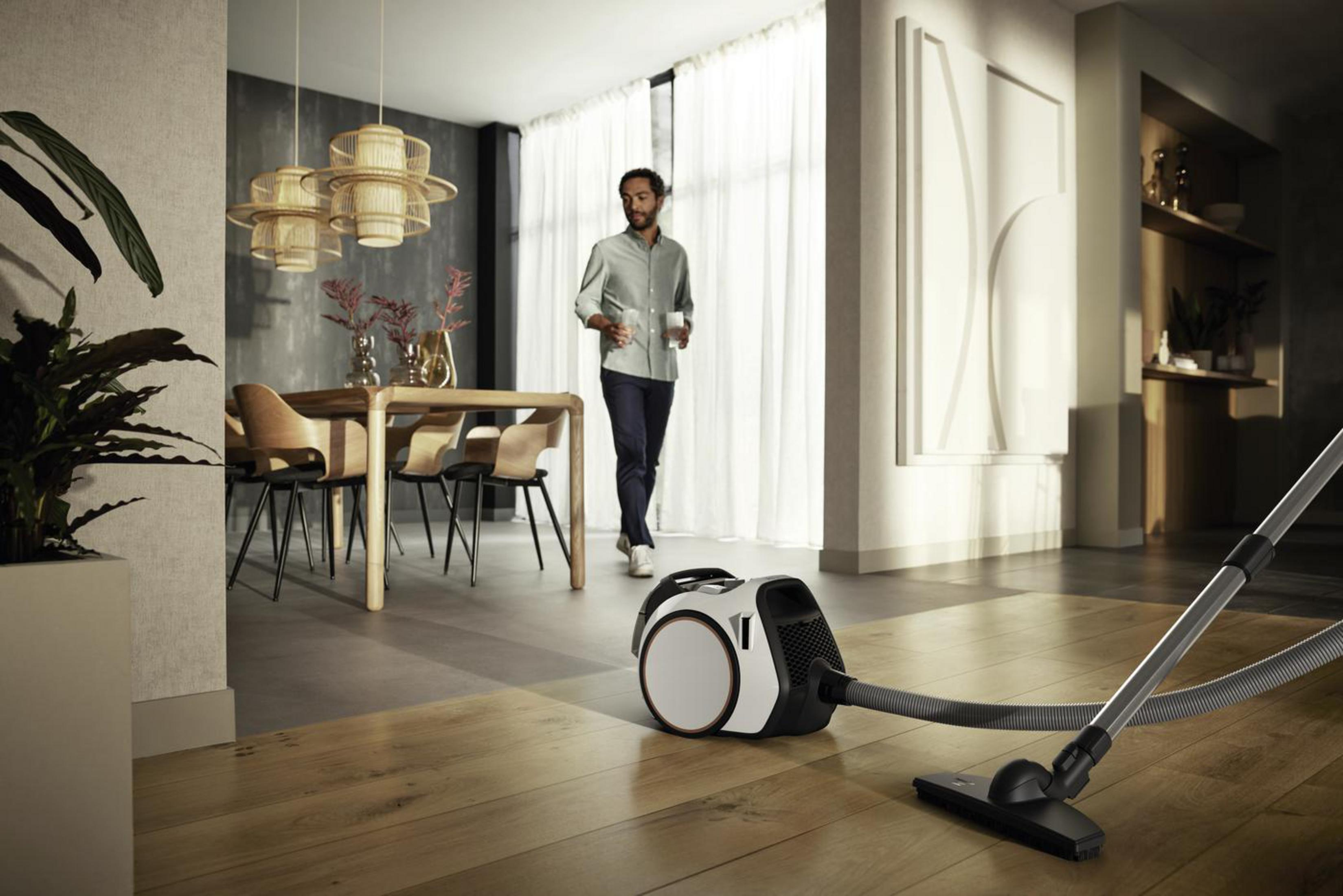 Scena w salonie. Mężczyzna przechodzi obok. Odkurzacz na pierwszym planie, stół z krzesłami.