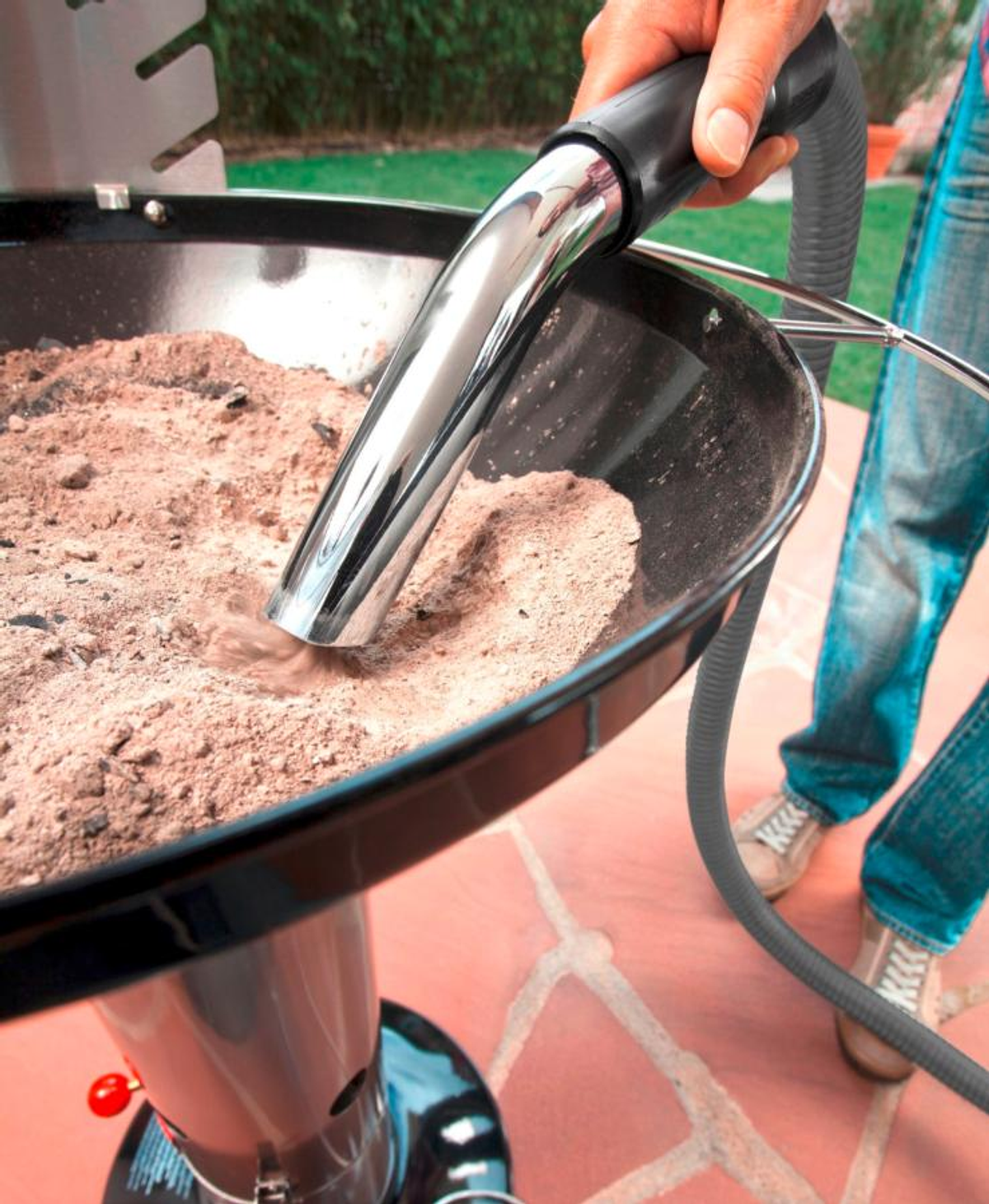 Osoba odkurzająca popiół z grilla za pomocą metalowej dyszy odkurzacza.