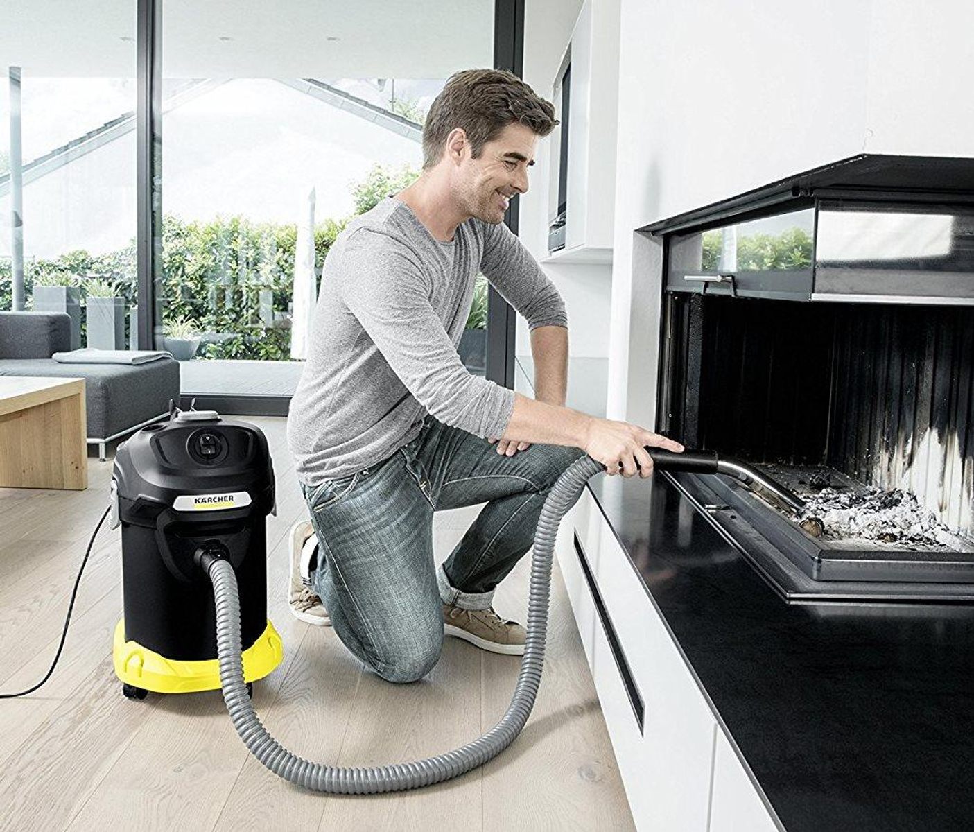 Mężczyzna czyści kominek odkurzaczem. Klęczy, ma szarą koszulę i dżinsy. Czarno-żółty odkurzacz.