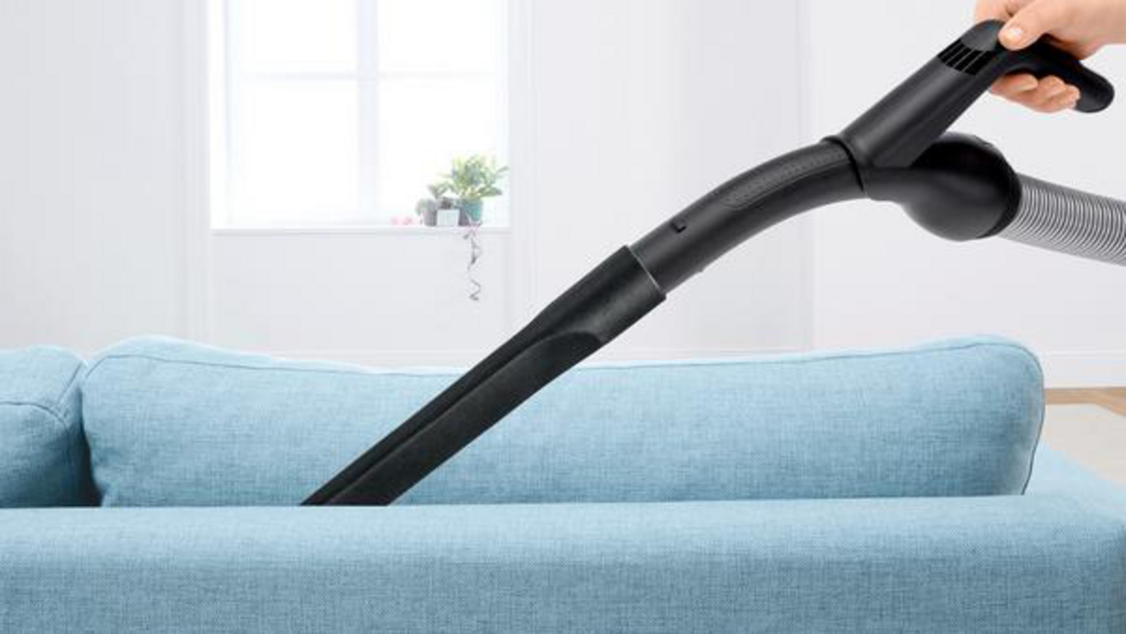 Staubsaugerdüse reinigt hellblaues Sofa. Hand hält den Griff. Hintergrund enthält Fenster.