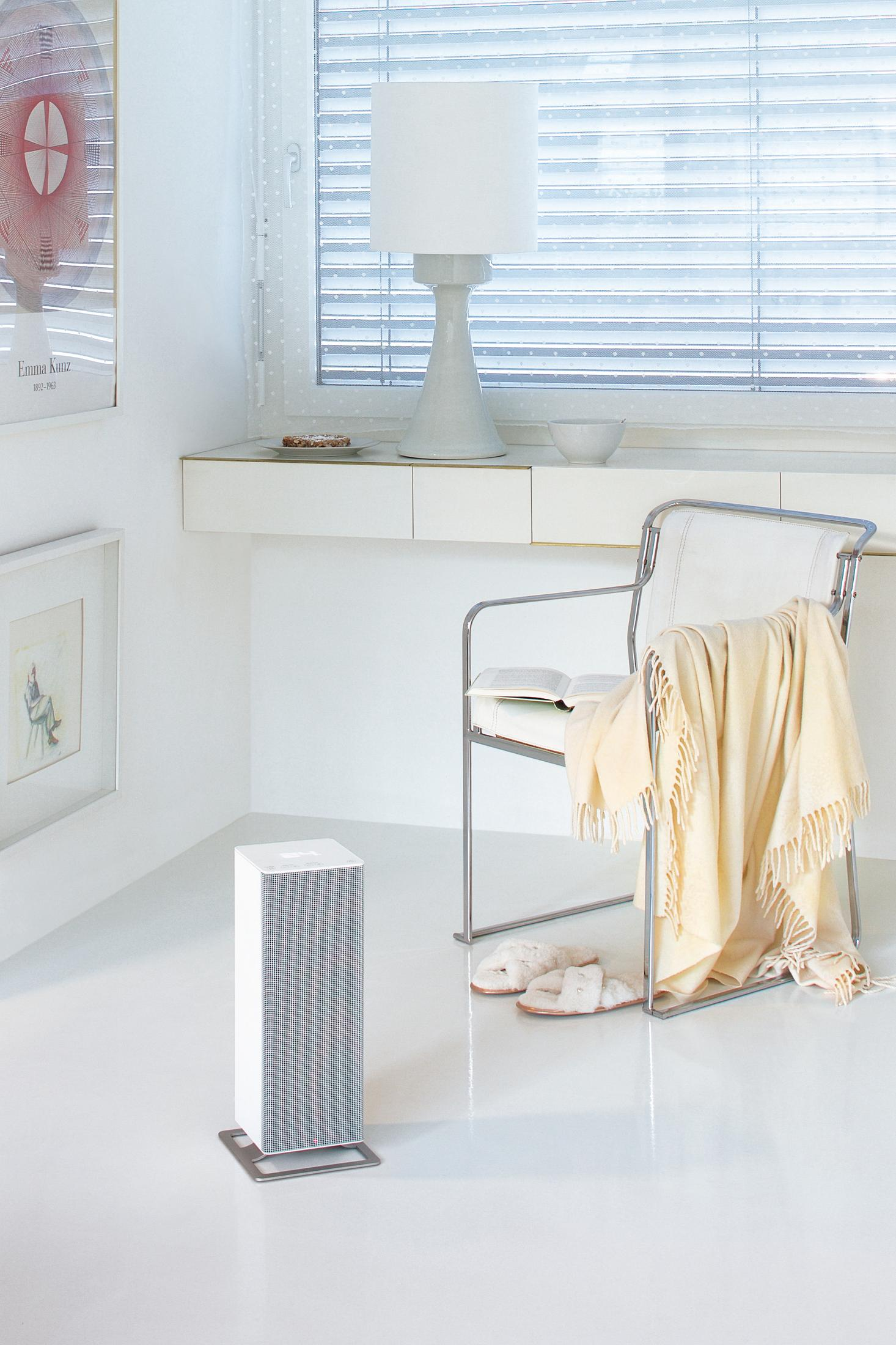Pièce blanche, avec un radiateur blanc, une chaise avec une couverture, une lampe et des pantoufles au sol.