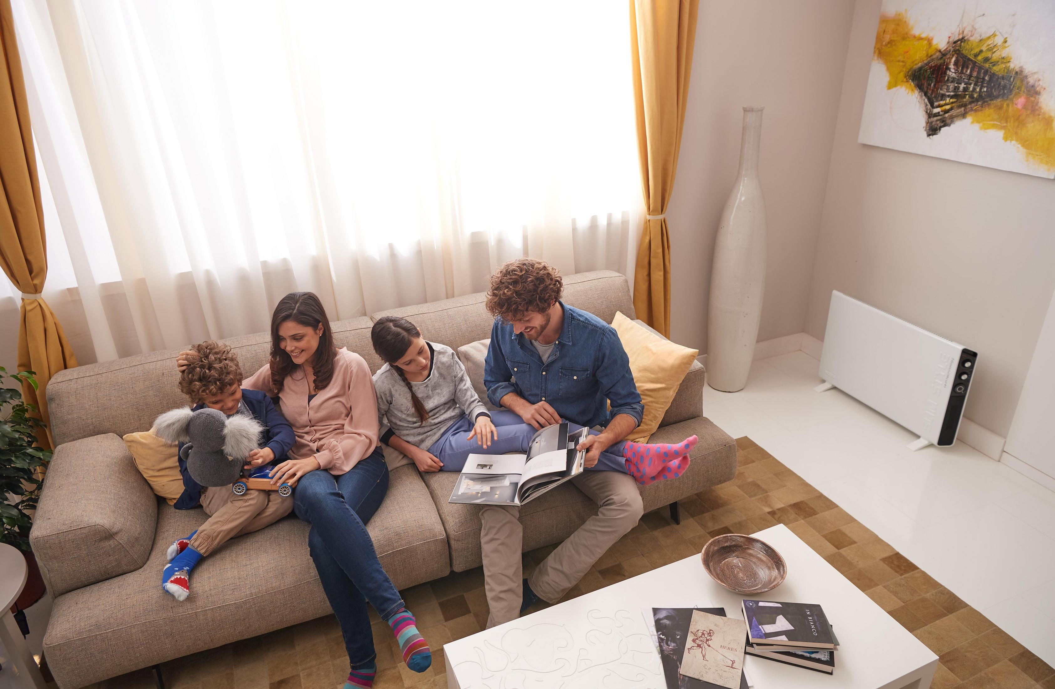Rodzina na sofie oglądająca album ze zdjęciami. Widoczna biała ściana, okno i grzejnik.