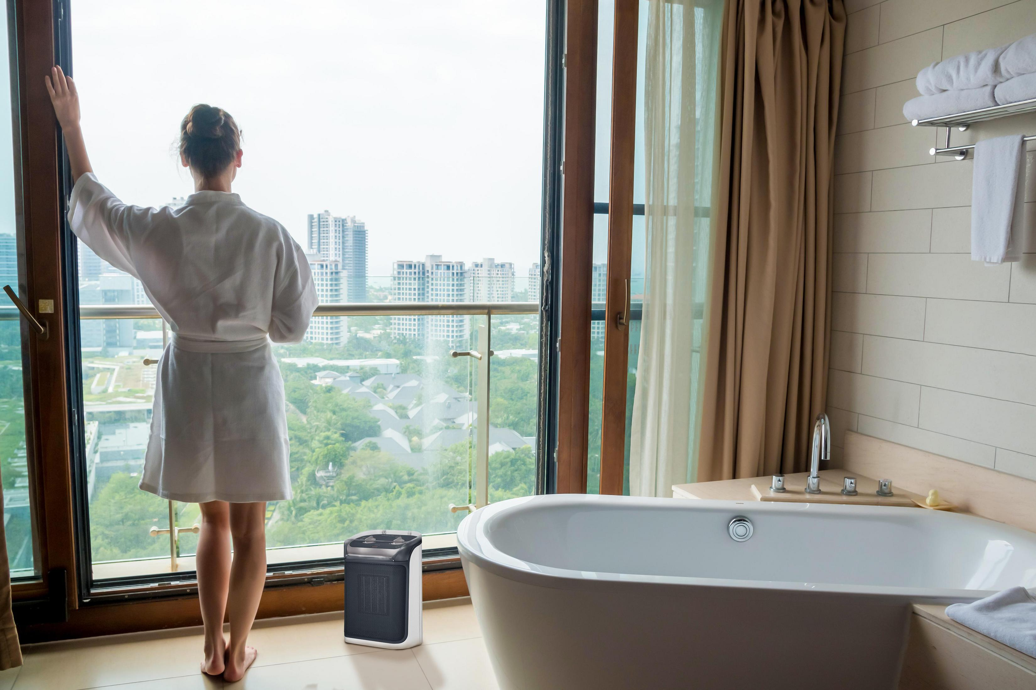 Kobieta w szlafroku stoi przy oknie, patrząc na widok miasta. Widoczna łazienka, wanna i grzejnik.
