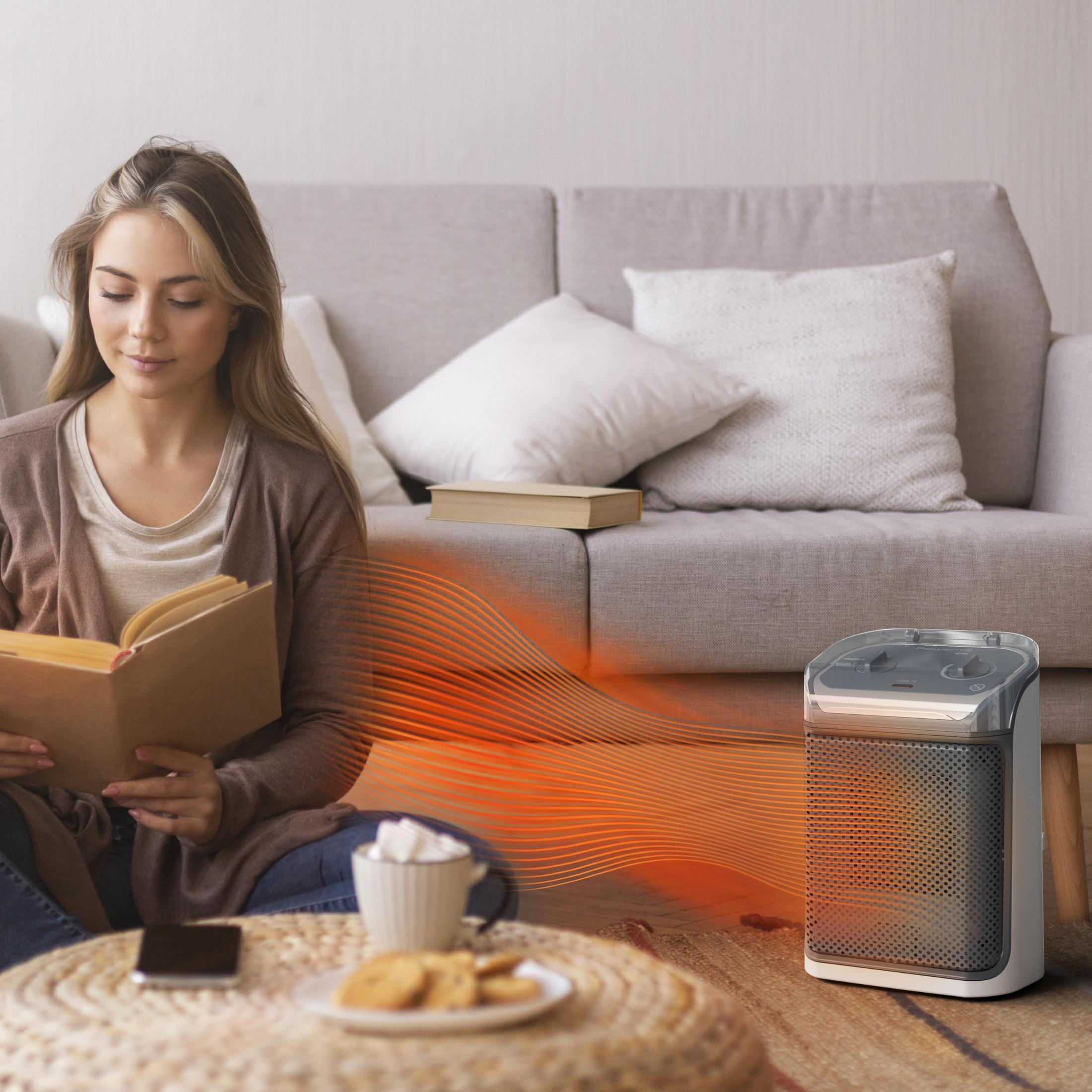 Kobieta czyta książkę, grzejnik na podłodze, kawa i ciastka na stole.