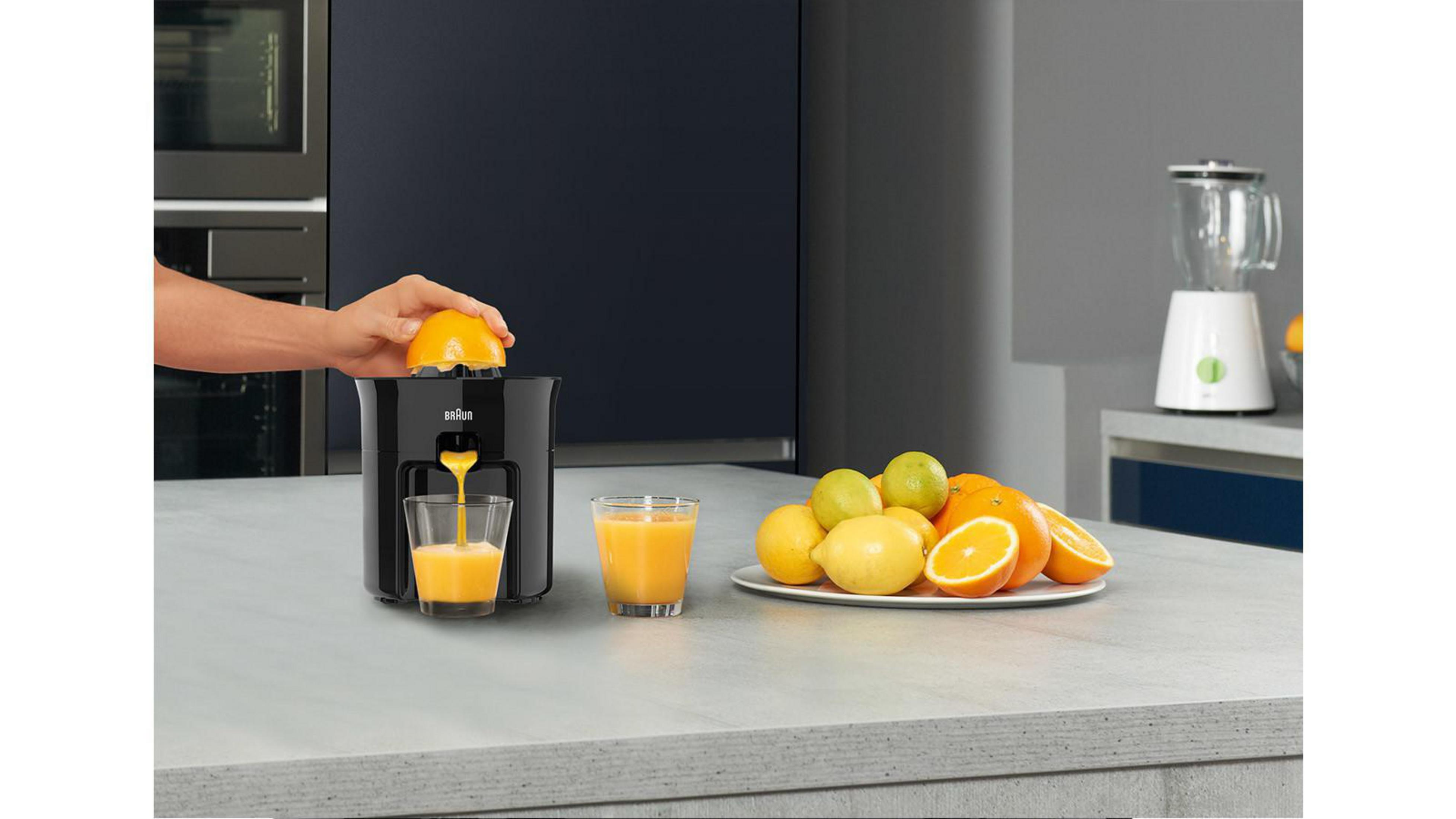 Osoba używająca czarnej wyciskarki. Pomarańcze i szklanki soku na blacie.