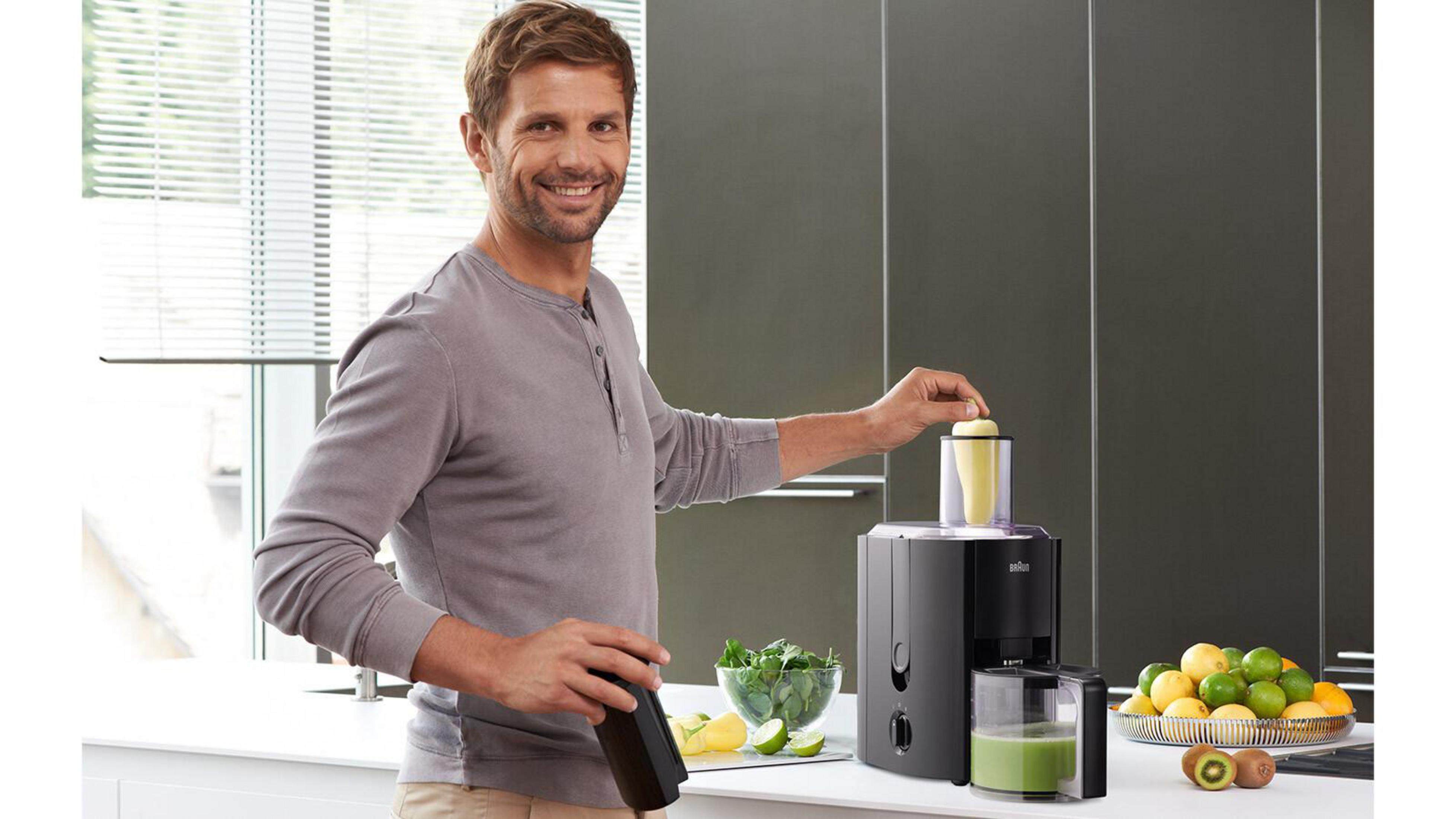 Mann macht Saft. Entsafter, Früchte und eine Schüssel mit Grünzeug auf der Theke.