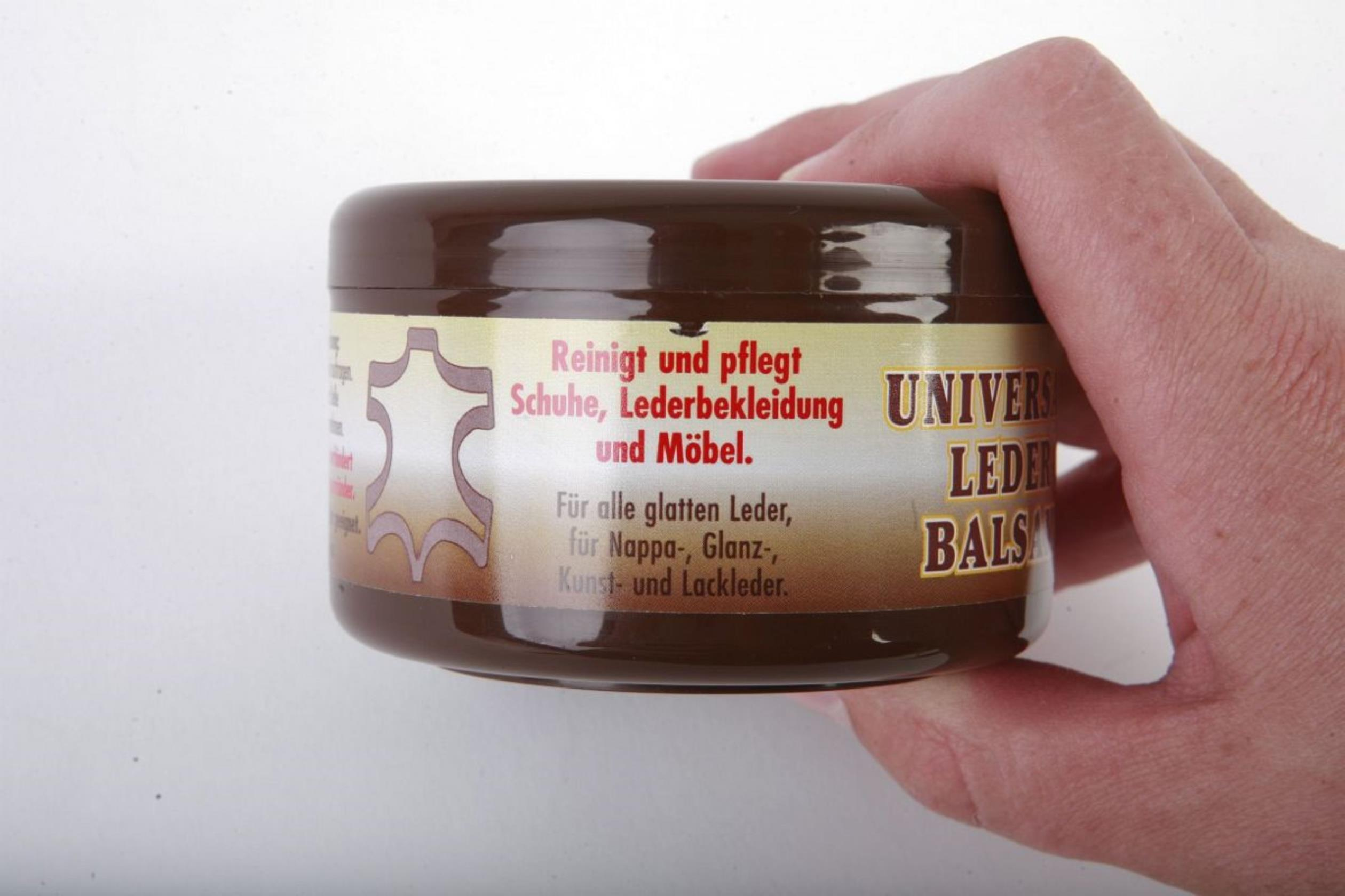 Ein braunes Glas Lederbalsam. Eine Hand hält das Glas. Textetiketten sind auf dem Glas sichtbar.