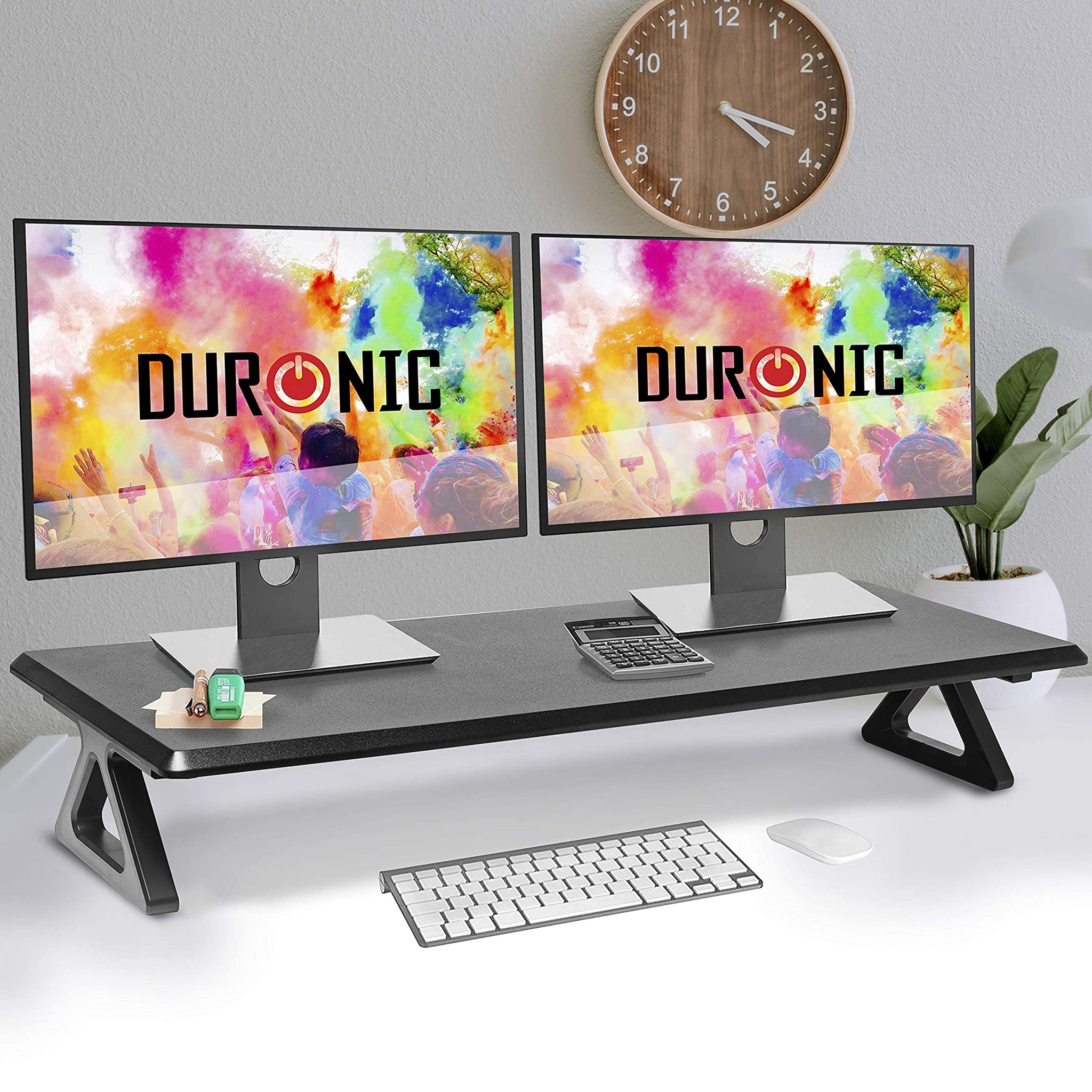 Zwei Monitore auf Schreibtischständer, Tastatur, Maus, Uhr und Pflanze.