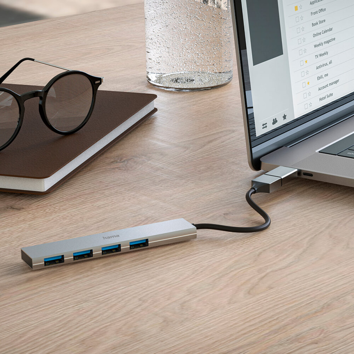 Laptop collegato a un hub USB argentato, occhiali e libro.