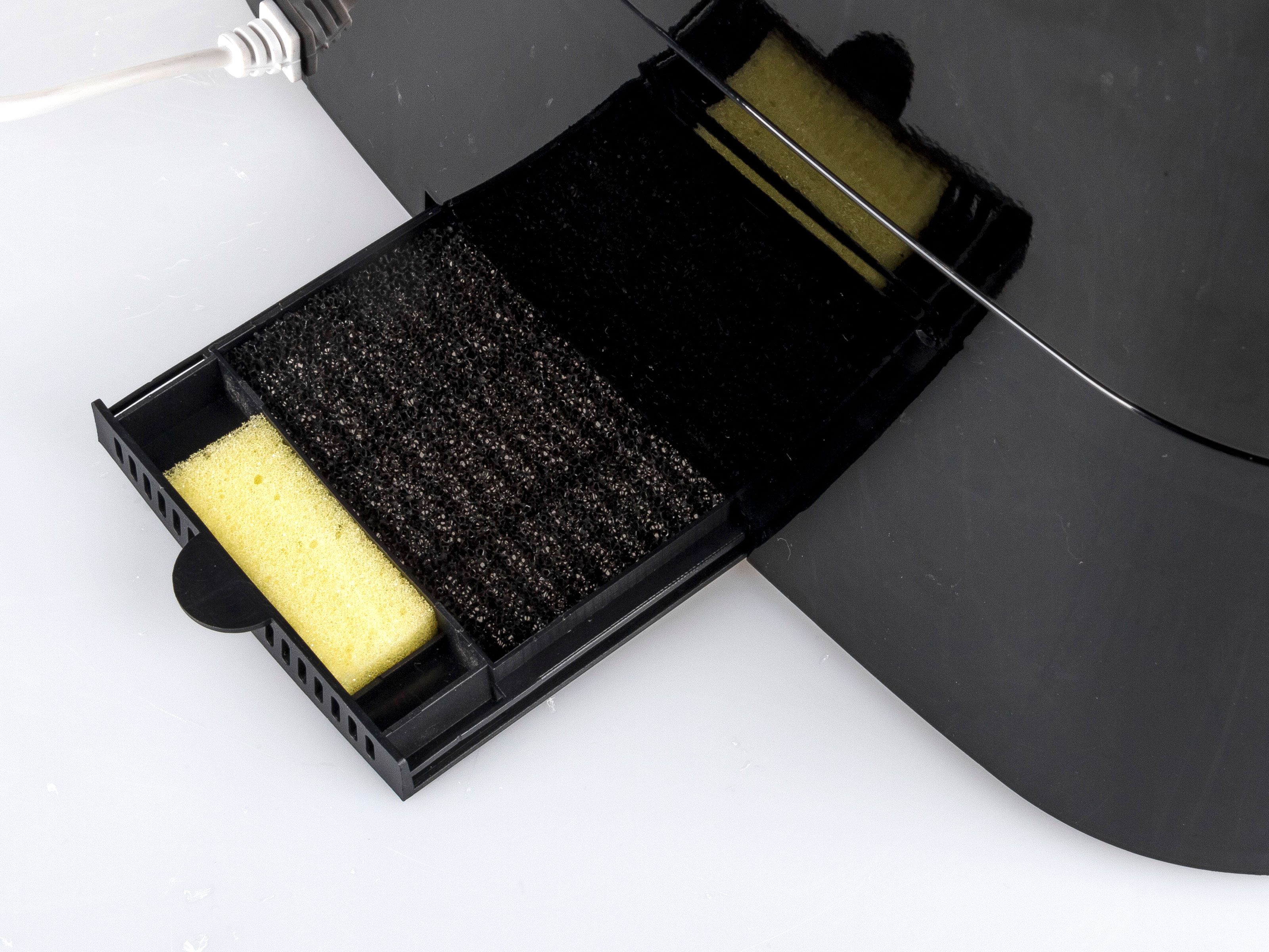 Ein schwarzer Filter mit einem gelben Schwamm und Filtermaterial im Inneren, auf weißer Oberfläche.