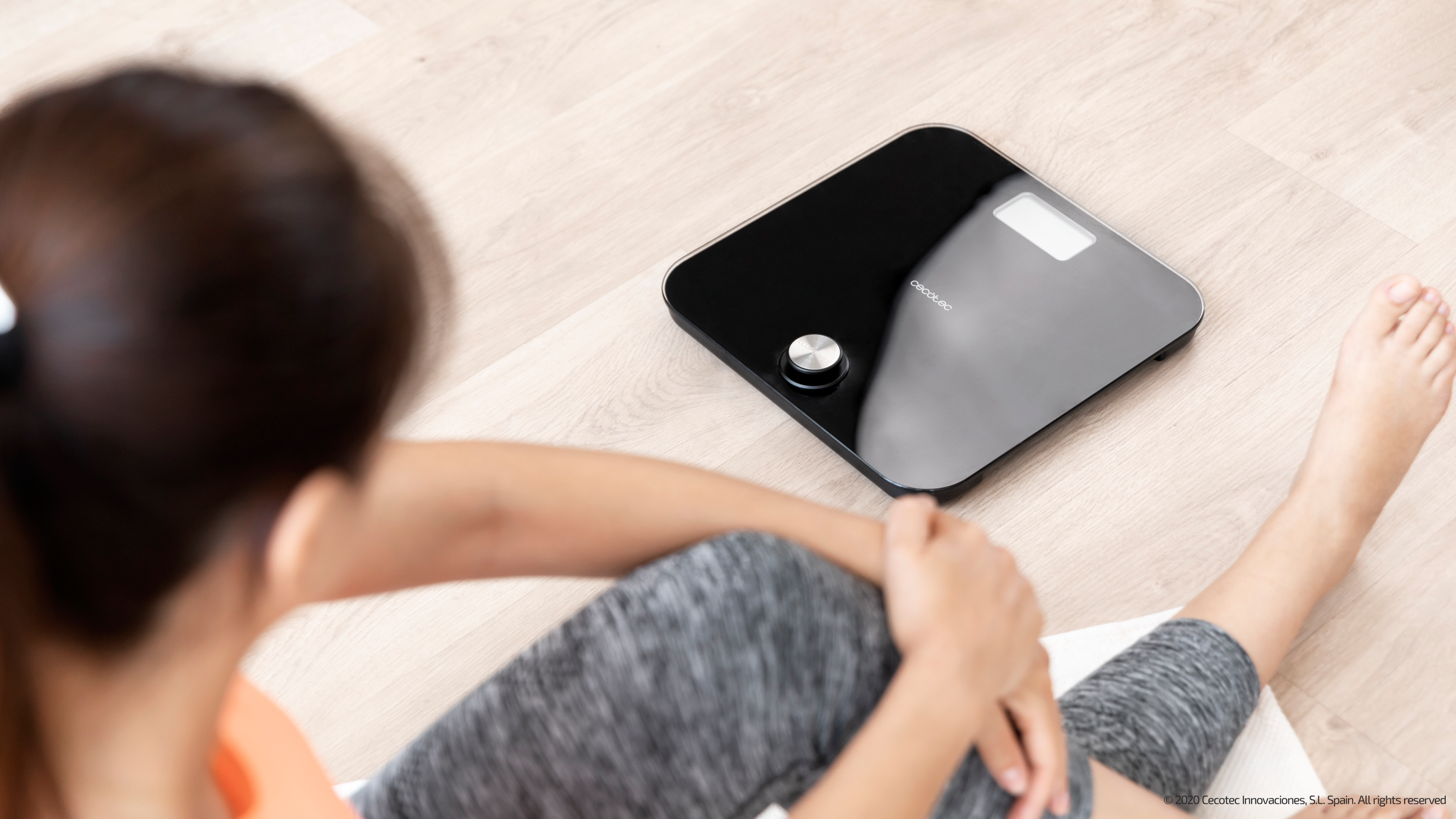 Kobieta siedzi obok czarnej wagi cyfrowej. Widoczne nogi, ubrana w strój do ćwiczeń.