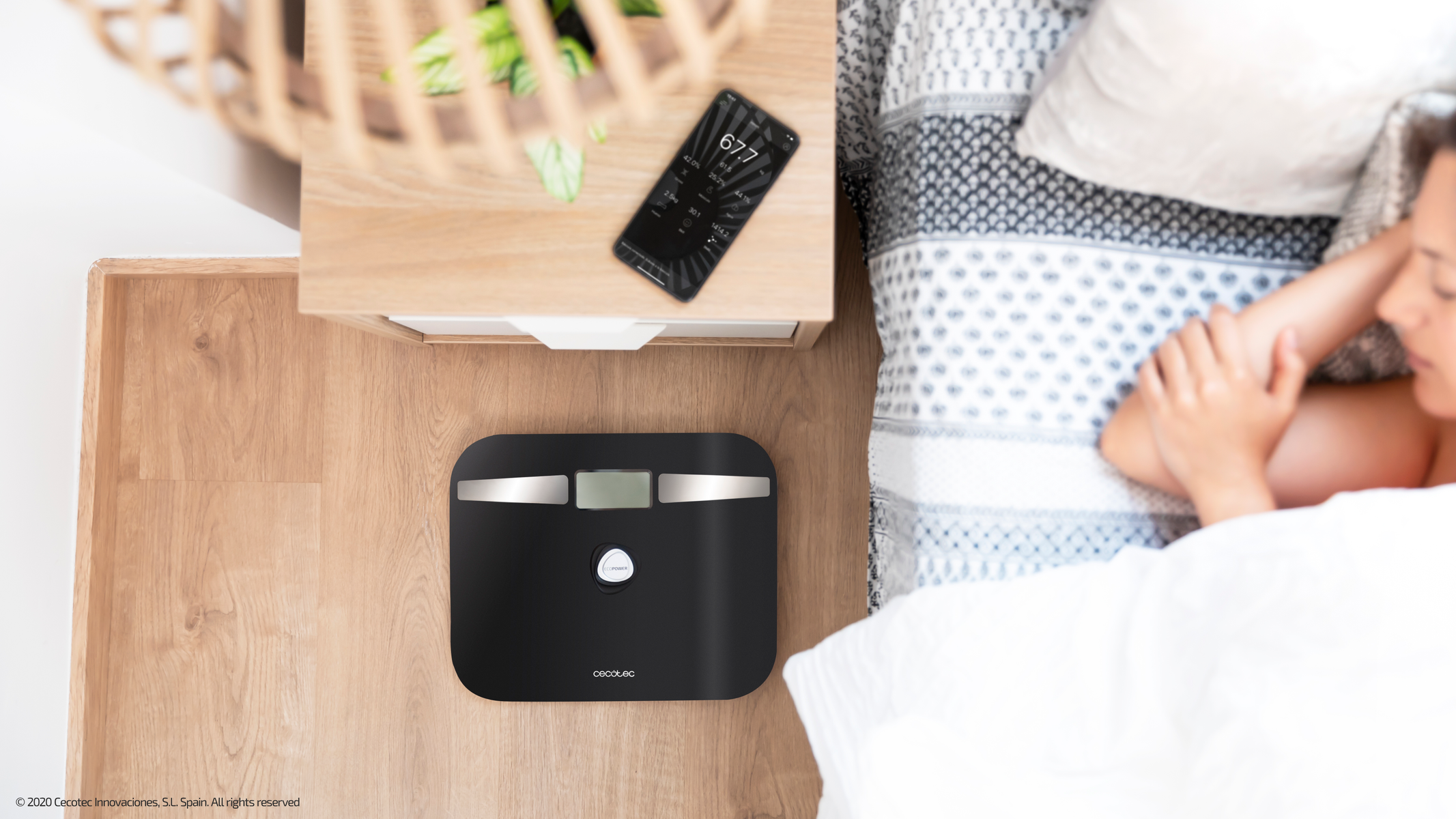 Czarna inteligentna waga obok łóżka ze śpiącą osobą. Widoczna szafka nocna z telefonem.