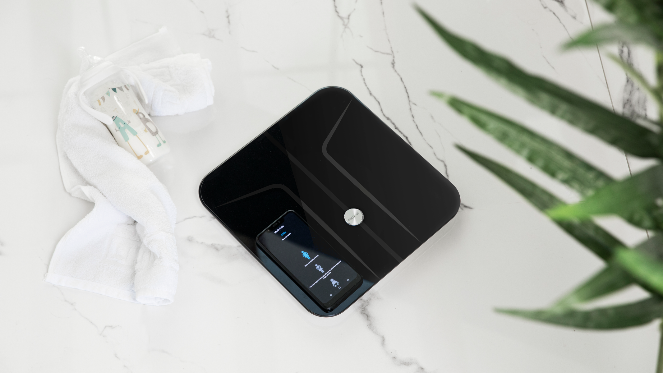 Czarna inteligentna waga ze smartfonem, ręcznikiem i kubkiem na białej marmurowej powierzchni.