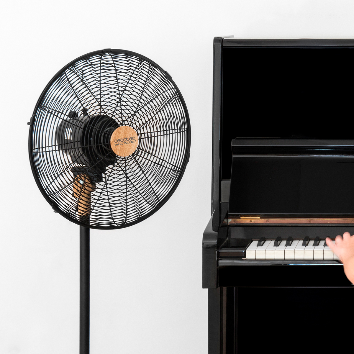 Czarny wentylator na statywie po lewej stronie czarnego fortepianu. Ręka przy klawiszach.