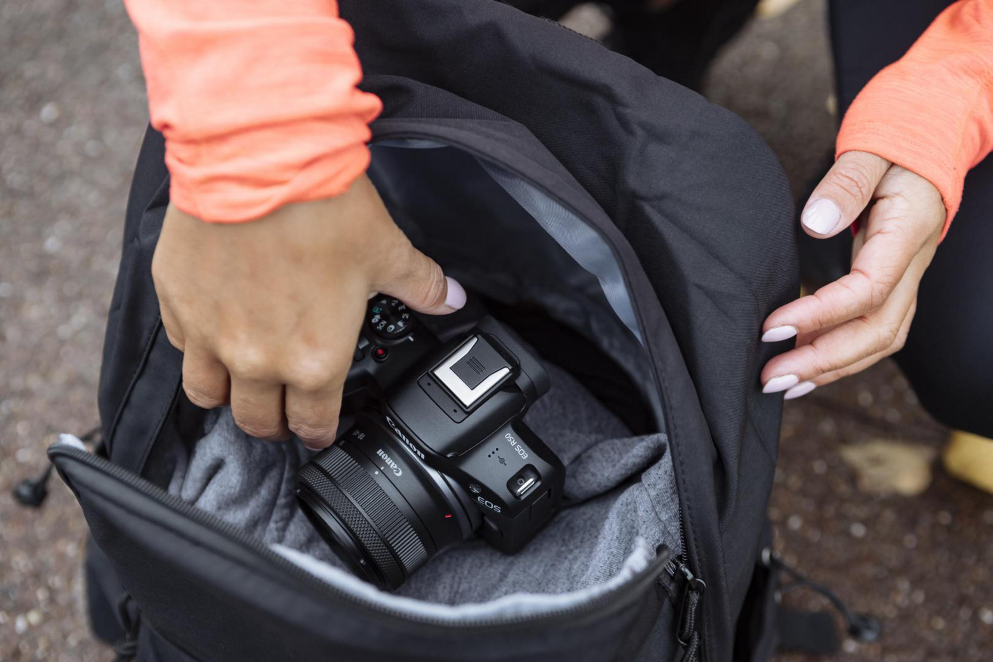 Hände legen eine Canon-Kamera in einen schwarzen Rucksack. Die Kamera ist schwarz mit Objektiv.