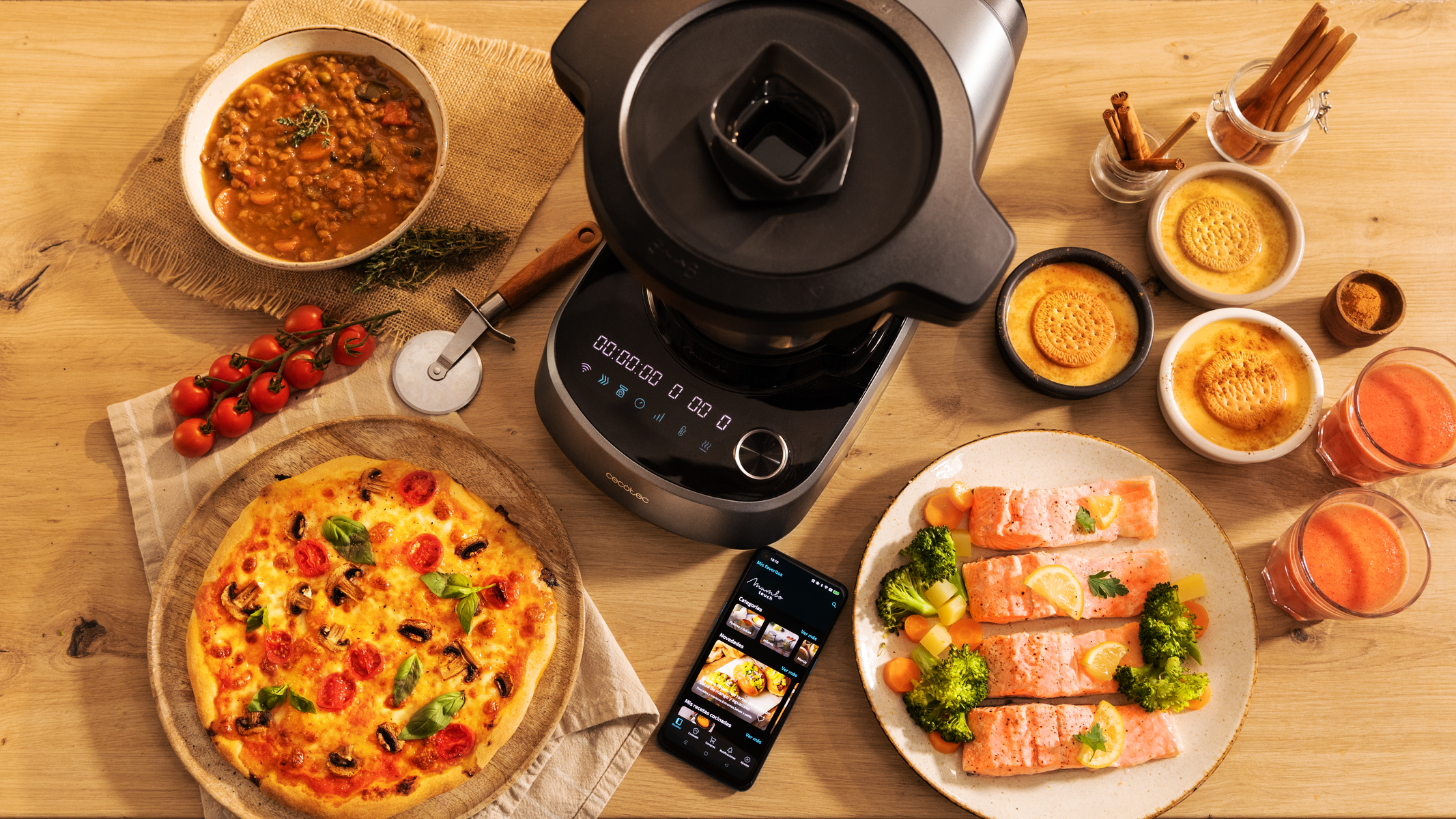 Vista cenital de una mesa con comida: pizza, salmón, sopa de lentejas y postres. Un teléfono muestra una receta.
