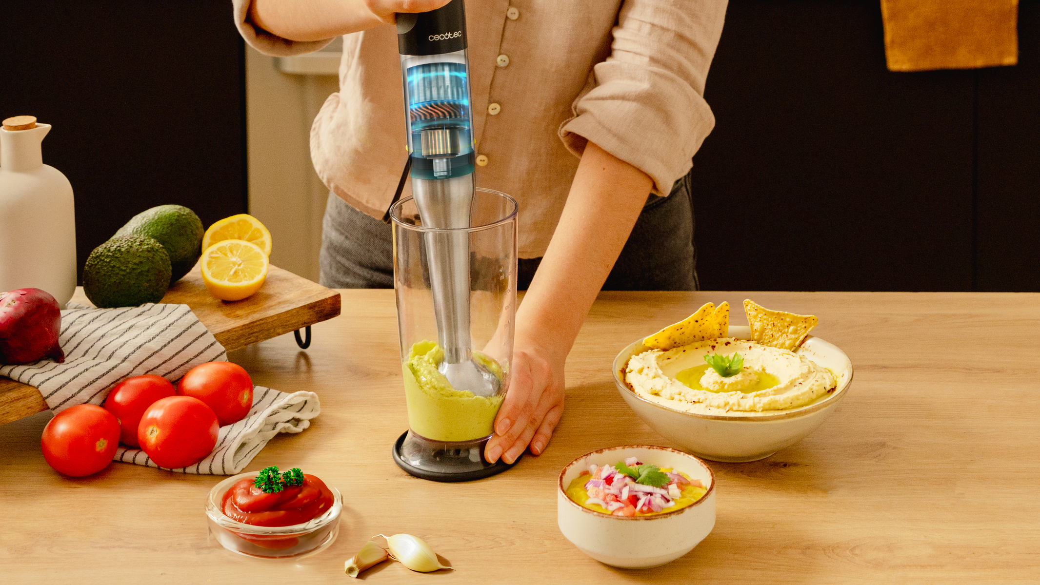 Osoba miksująca awokado blenderem ręcznym w szklance. Warzywa i dipy na drewnianym stole.