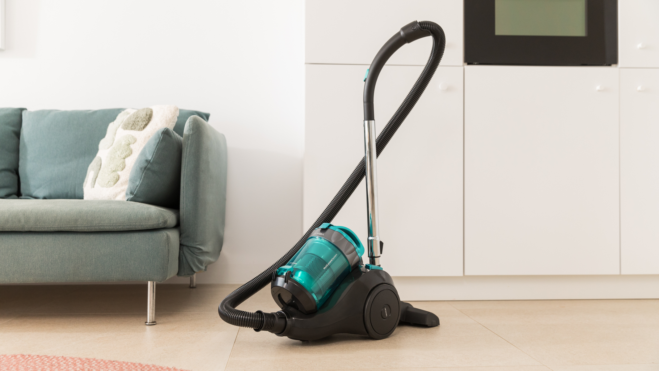 Odkurzacz na drewnianej podłodze w białym pokoju. Turkusowa sofa w tle.