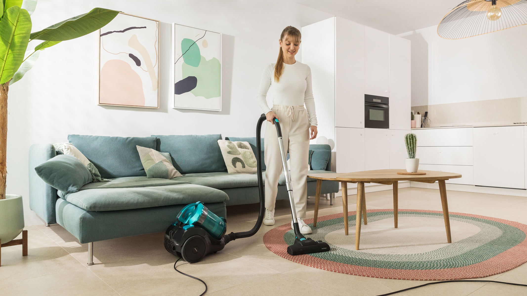 Kobieta odkurza salon. Widoczna turkusowa kanapa, sztuka i dywan. Białe szafki kuchenne w tle.