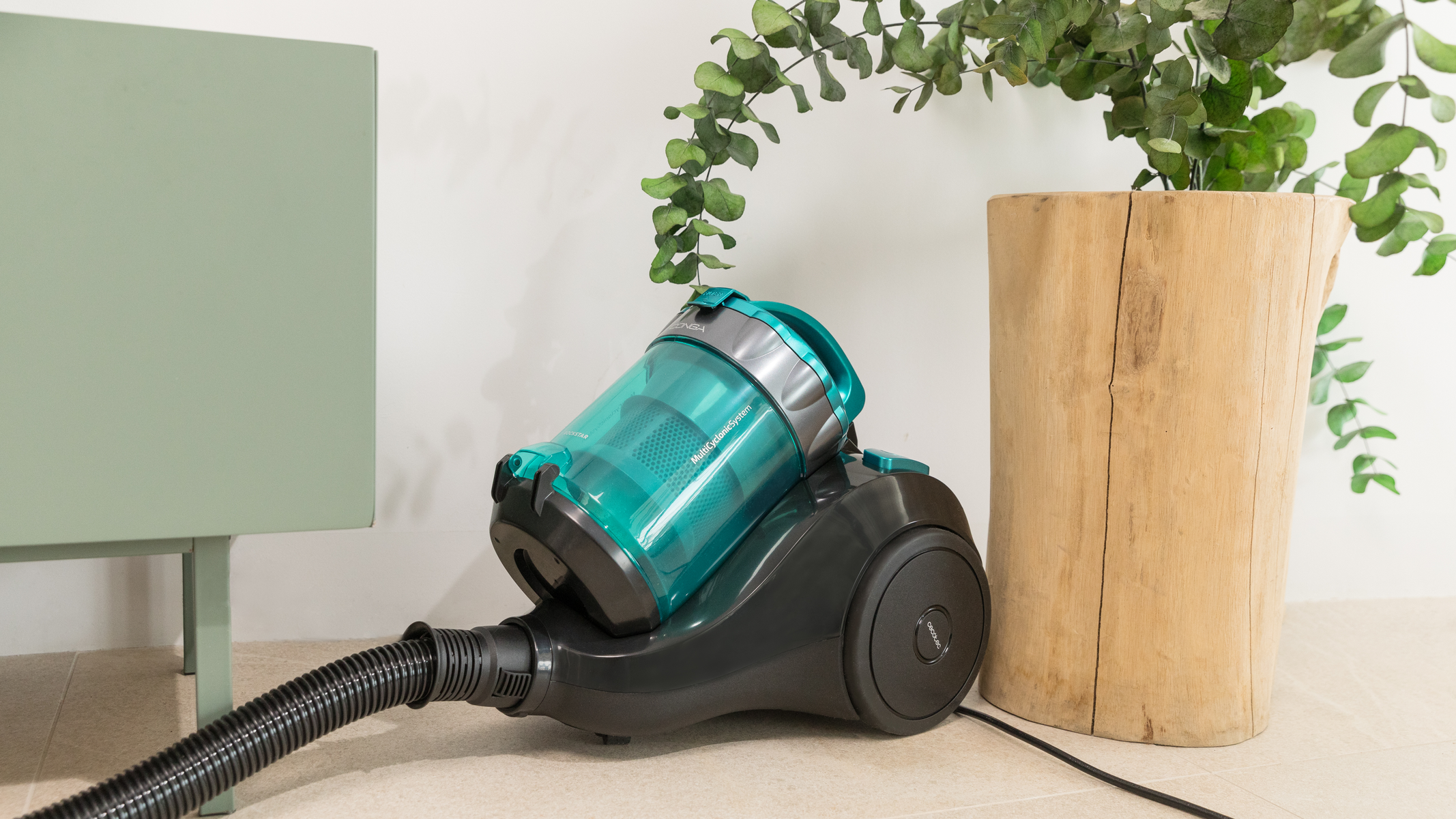 Turkusowy i czarny odkurzacz z wężem, na podłodze obok drewnianego pnia.