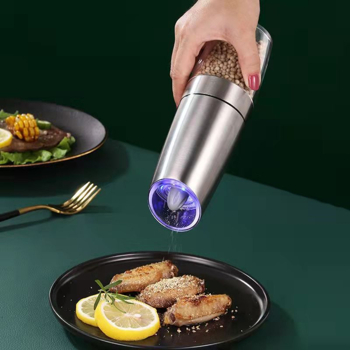 Eine Hand mahlt Pfeffer über Essen. Ein Teller mit gebratenen Hähnchenflügeln und Zitronenscheiben ist sichtbar.