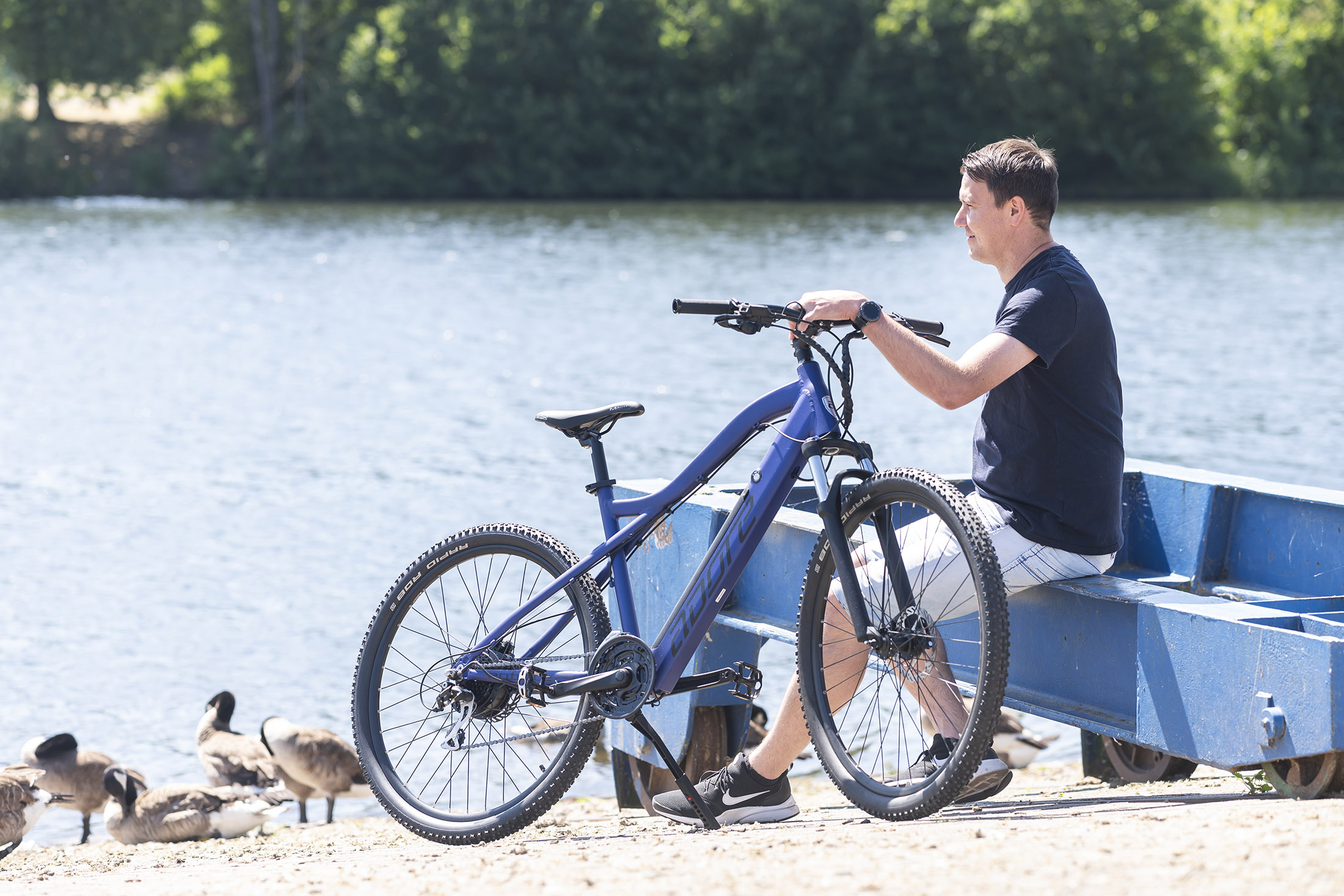 Ein Mann sitzt an einem See neben einem blauen Fahrrad. Gänse schwimmen in der Nähe im Wasser.