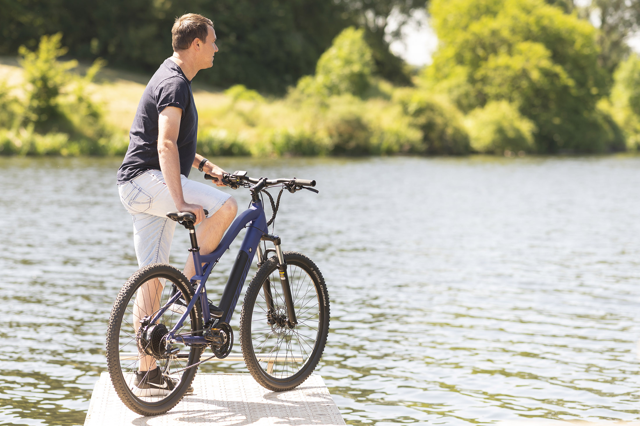 Ein Mann steht auf einem Steg neben einem blauen Elektrofahrrad und blickt auf einen See.