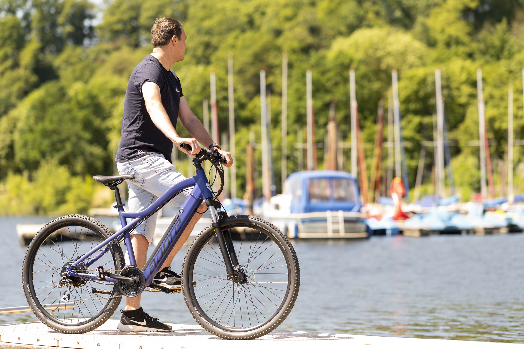 Mann mit Fahrrad auf einem Steg, Wasser und Boote im Hintergrund.