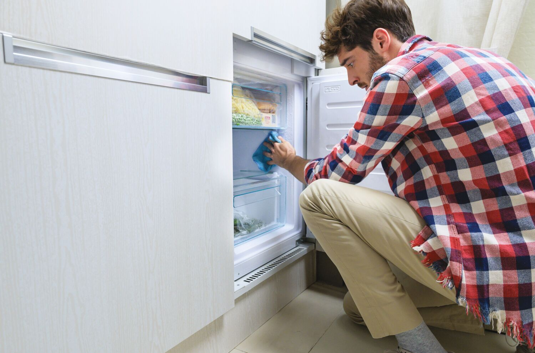 Un uomo pulisce un frigorifero, indossa una camicia a quadri e pantaloni color cachi. La porta del frigorifero è aperta.