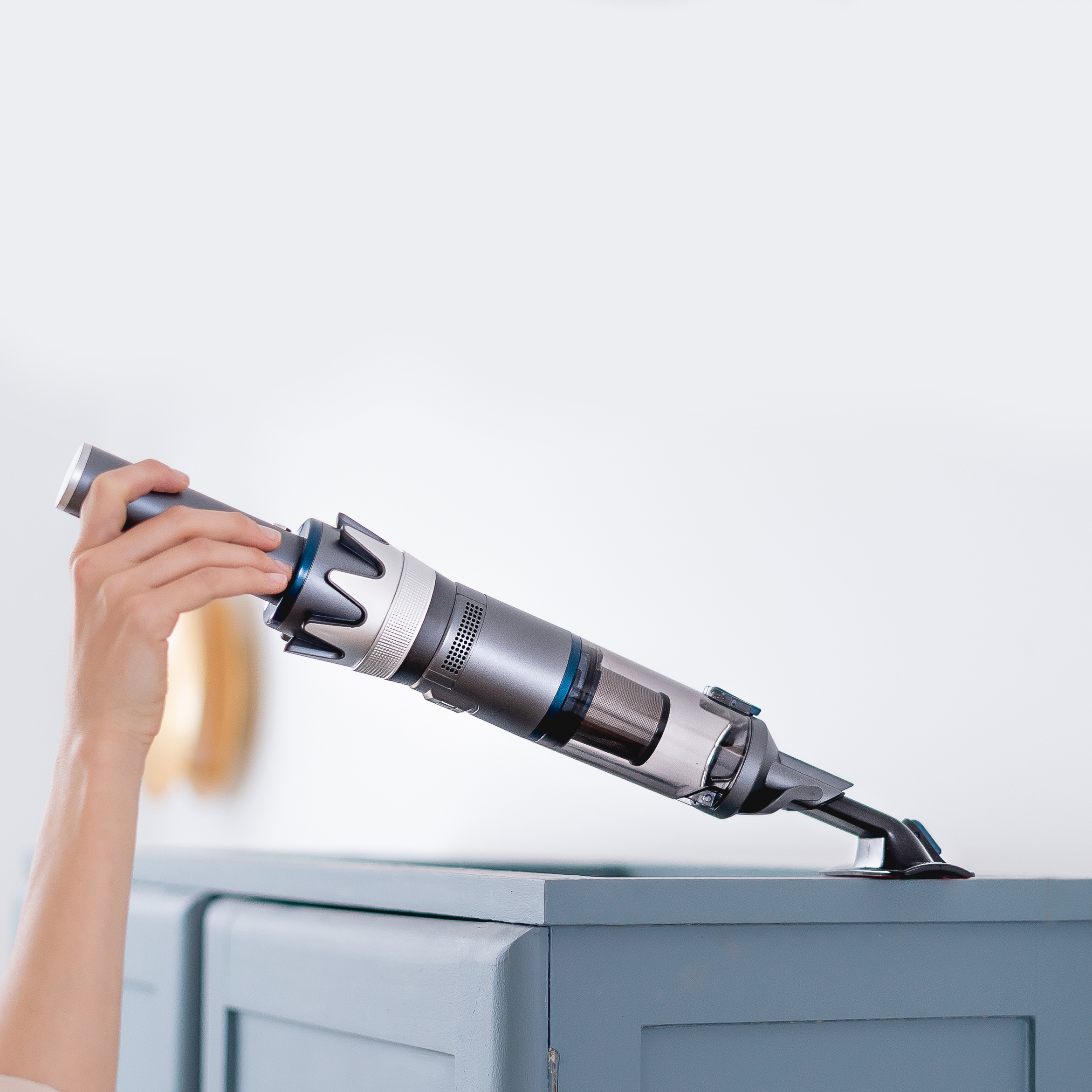Hand einer Person, die einen silbernen und grauen kabellosen Staubsauger über einem blauen Schrank hält.