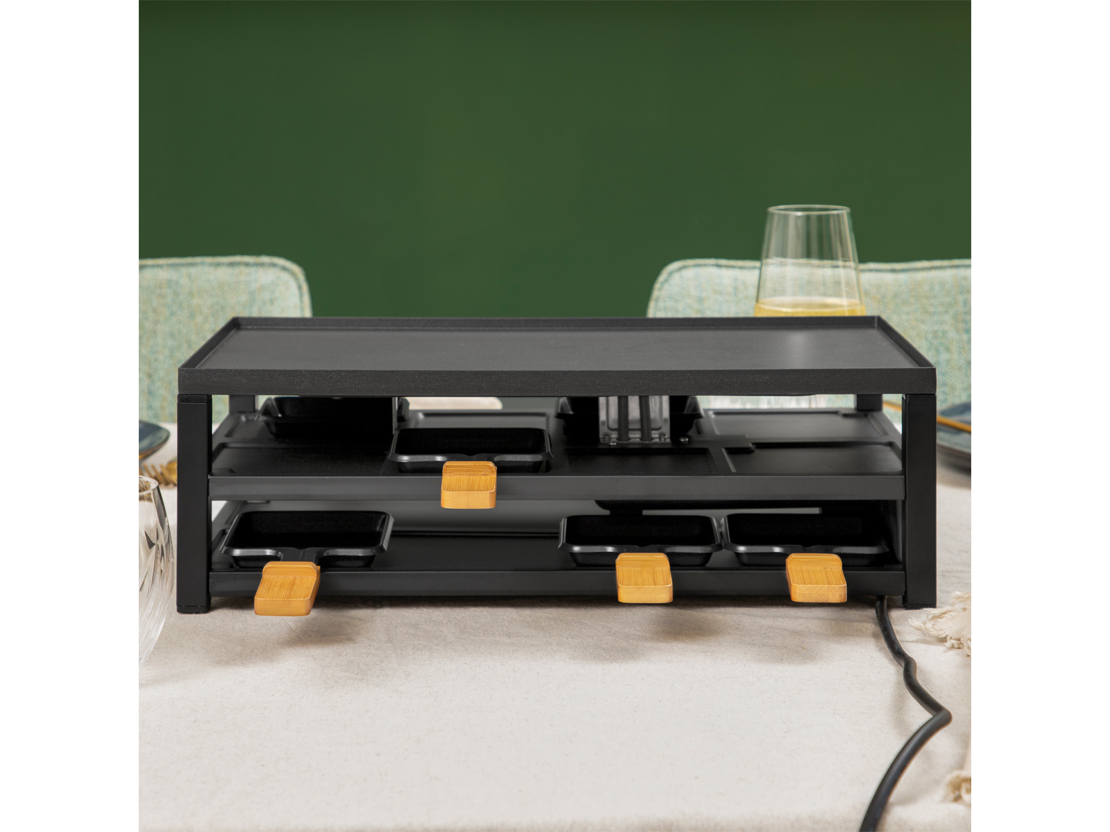 Czarny grill do raclette z wieloma patelniami i drewnianymi uchwytami, na stole. Szklanka płynu w tle.