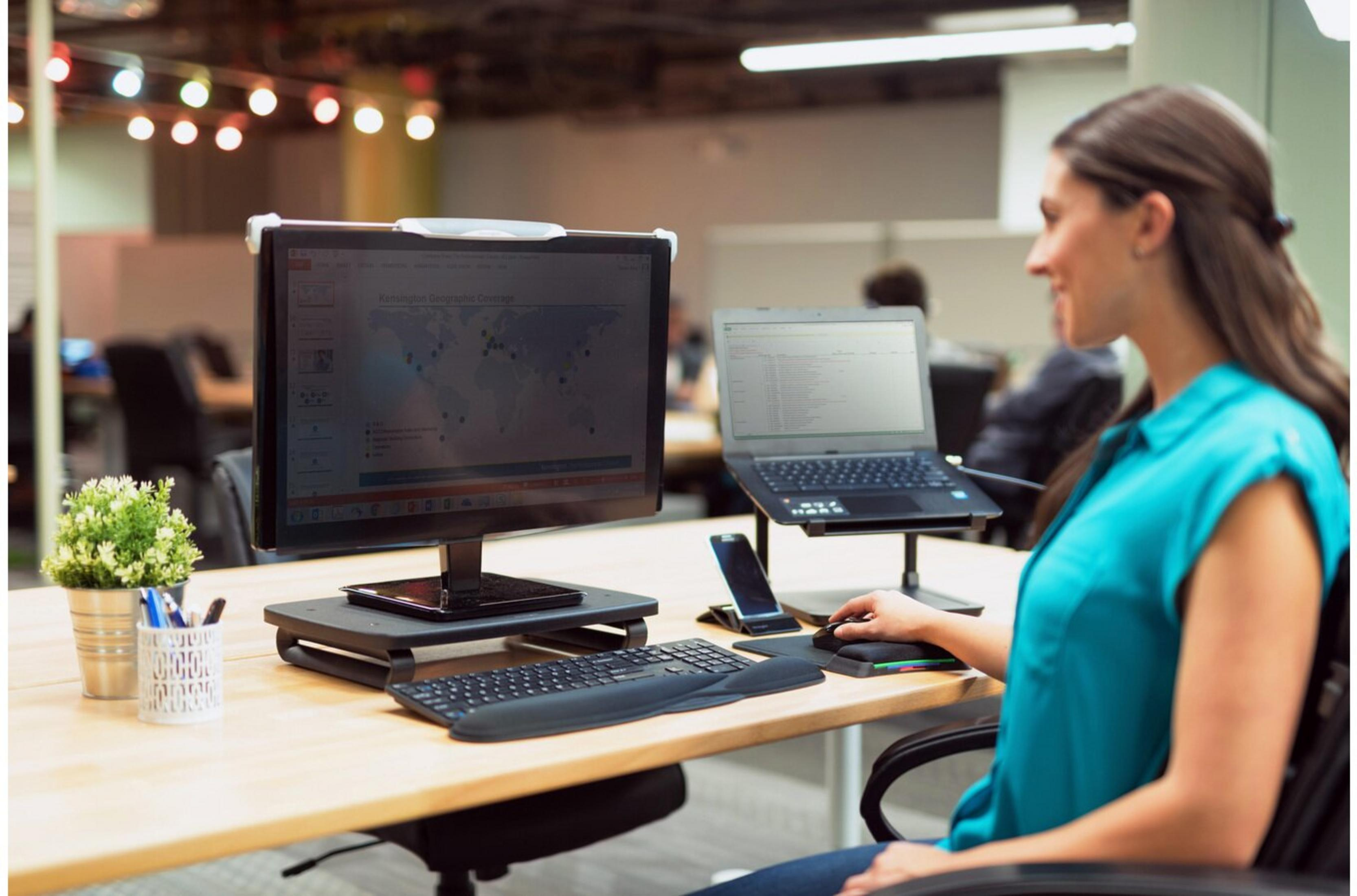 Eine Frau arbeitet an einem Schreibtisch mit Computer, Laptop und Telefon. Es gibt Pflanzen und Zubehör.