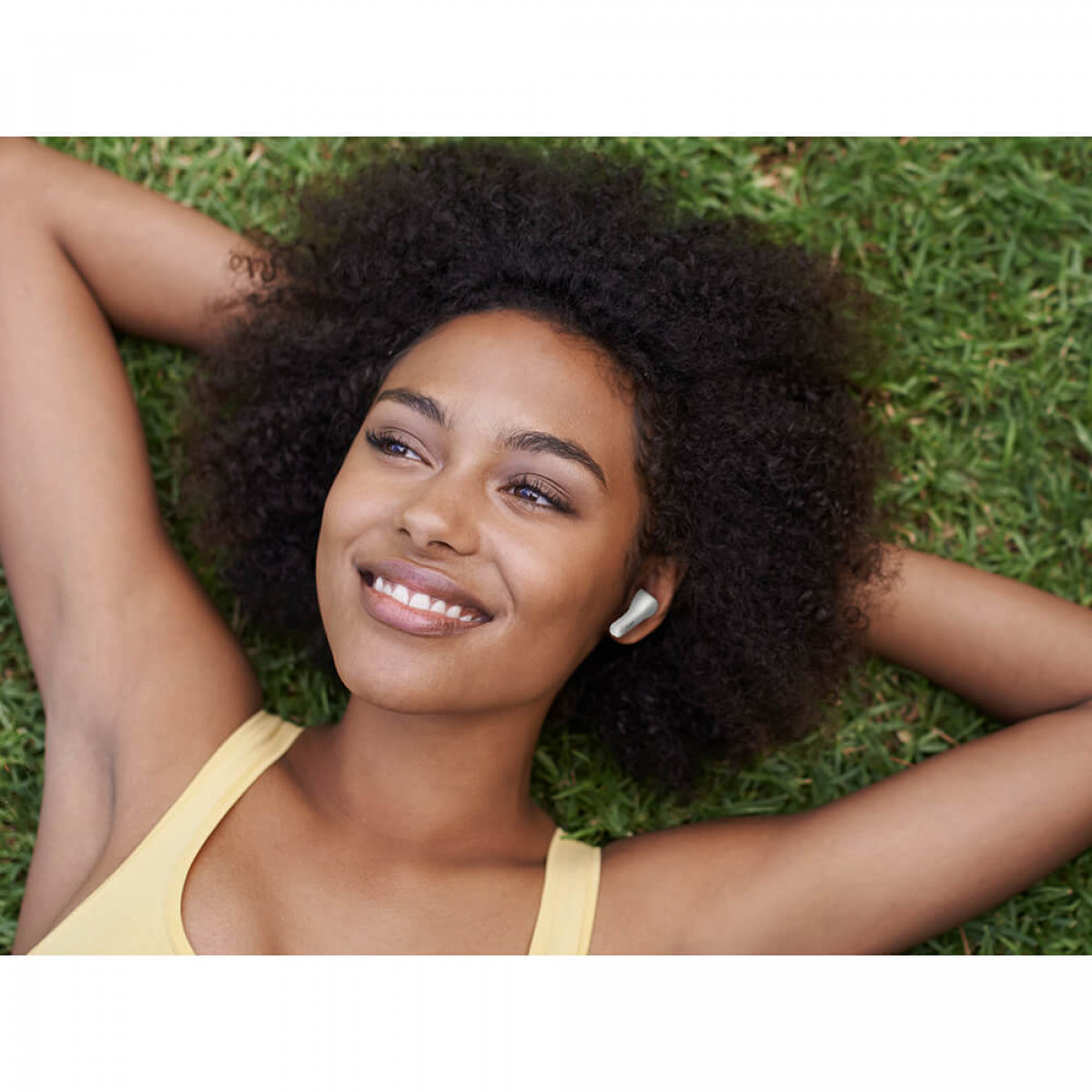 Kobieta w żółtym topie, leżąca na trawie ze słuchawkami, uśmiechnięta.