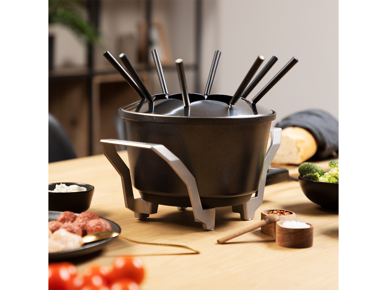 Zestaw do fondue z widelcami, czarny garnek na stojaku, otoczony składnikami na stole.