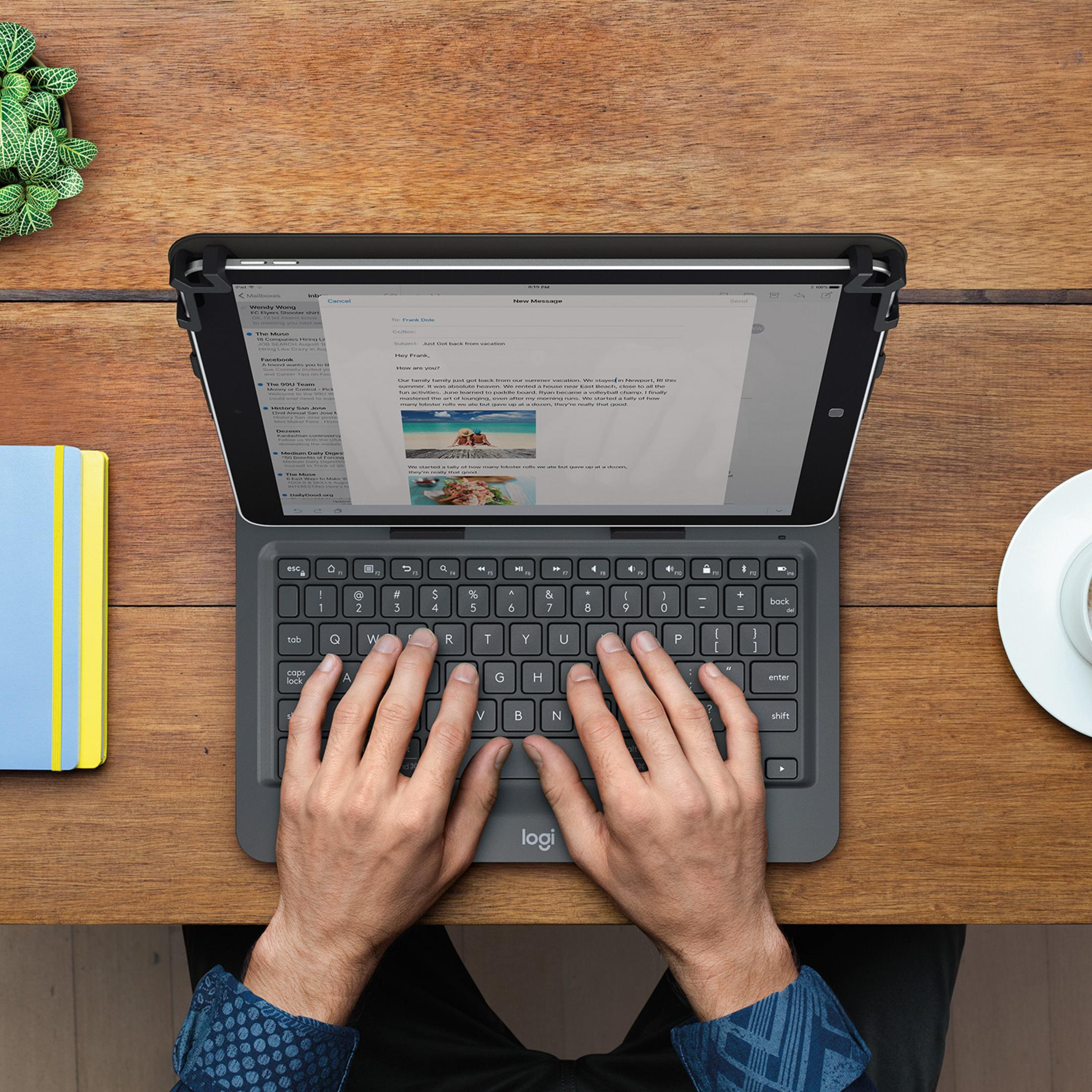 Eine Person tippt auf einer Tastatur, die an einem Tablet befestigt ist. Ein Notizbuch und eine Kaffeetasse stehen auf dem Tisch.