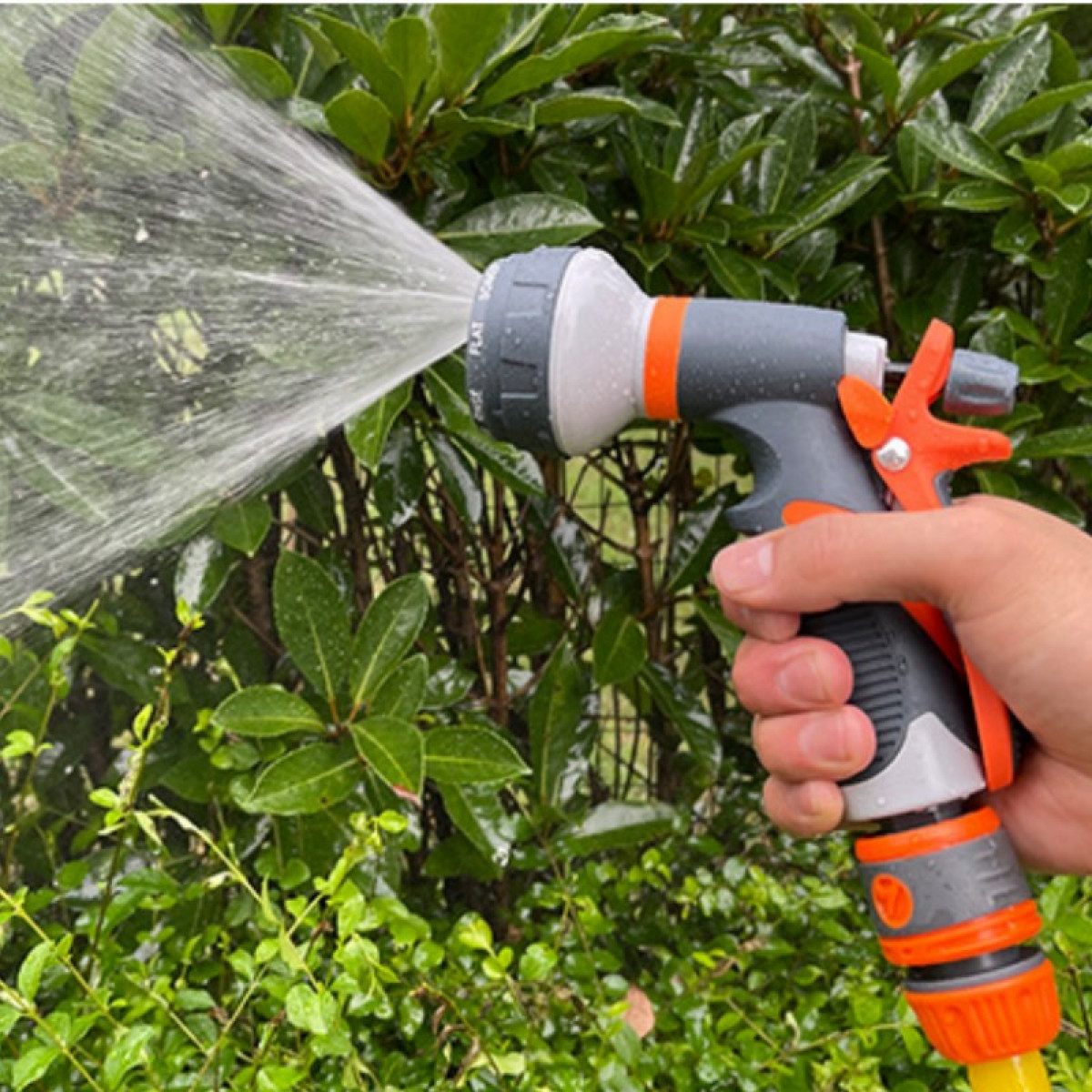 Hand hält graue und orange Wasserspritzpistole, sprüht Wasser auf Blätter.