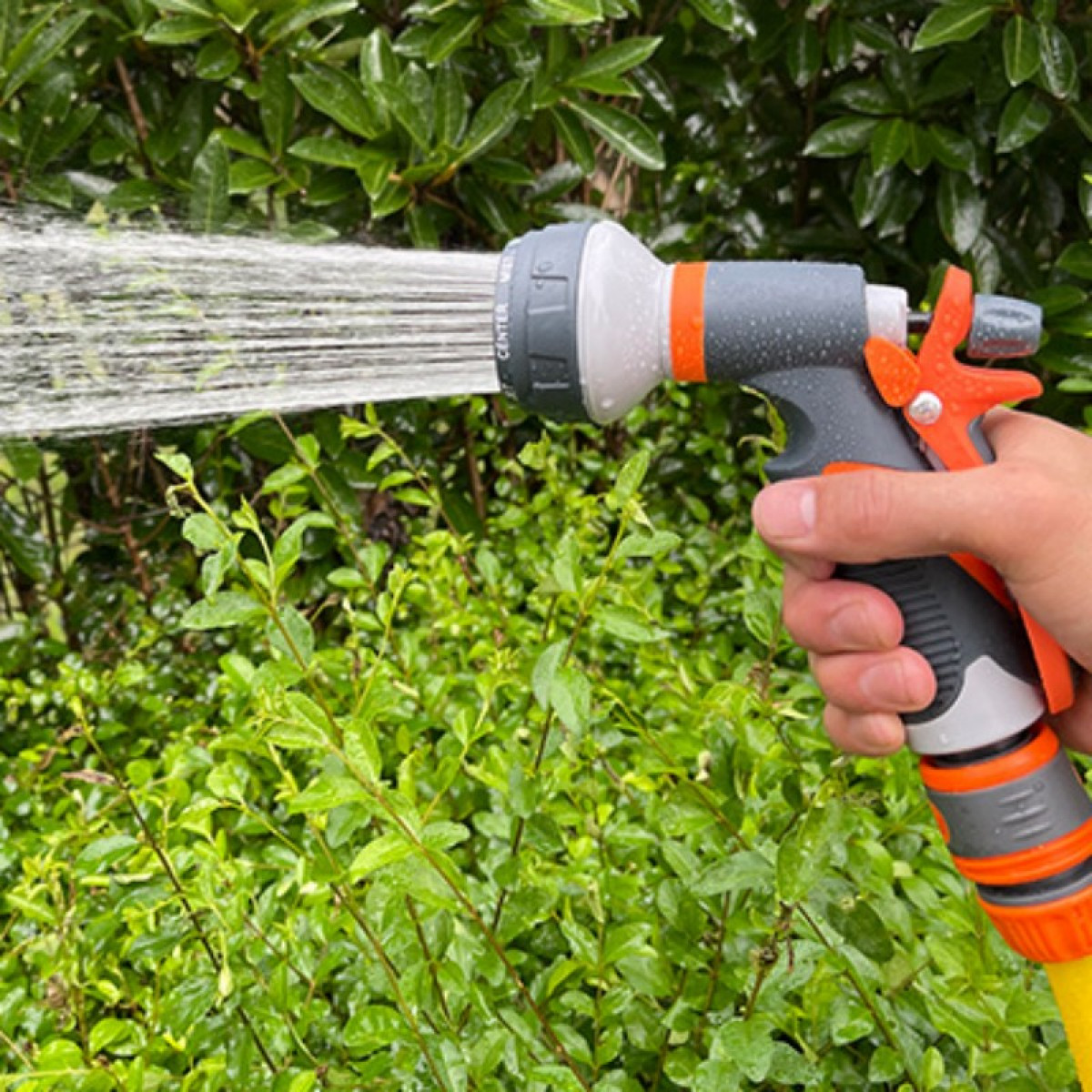 Eine Hand, die eine grau-orangefarbene Wasserschlauchdüse hält und Wasser auf grüne Büsche sprüht.