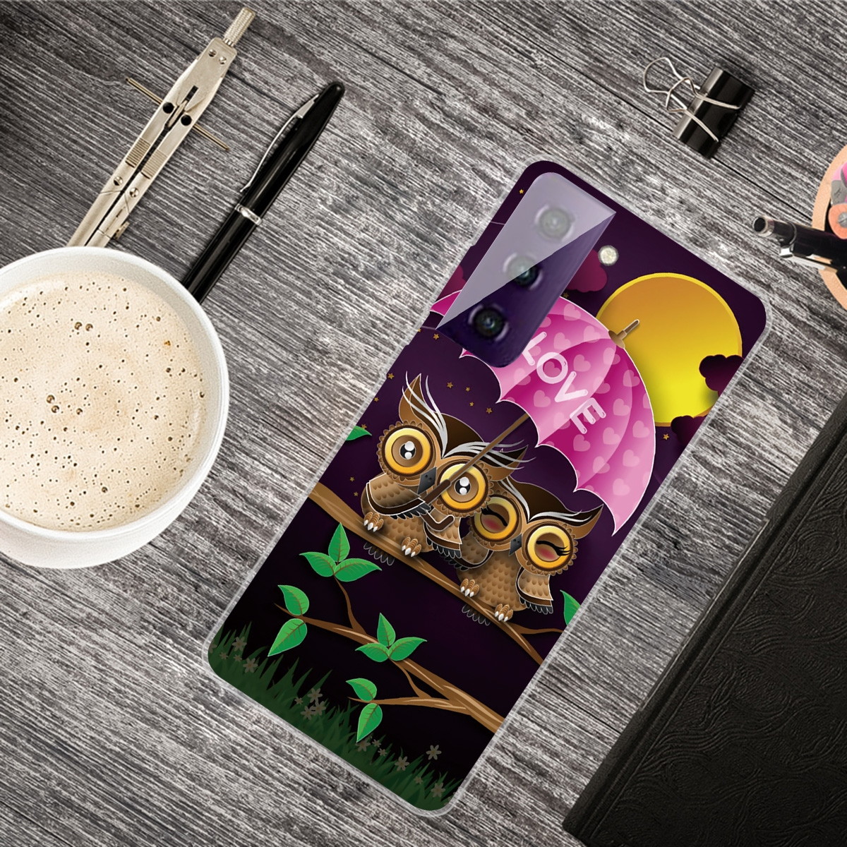 Funda de teléfono con búhos bajo un paraguas rosa, sobre un escritorio de madera con café y otros objetos.