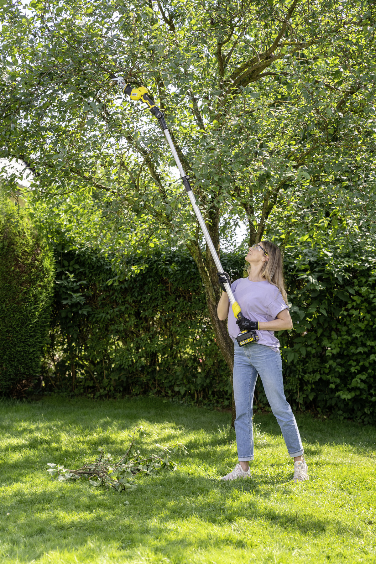 Eine Frau benutzt eine Stangensäge, um einen Baum in einem grünen Garten zu beschneiden, sonniger Tag.