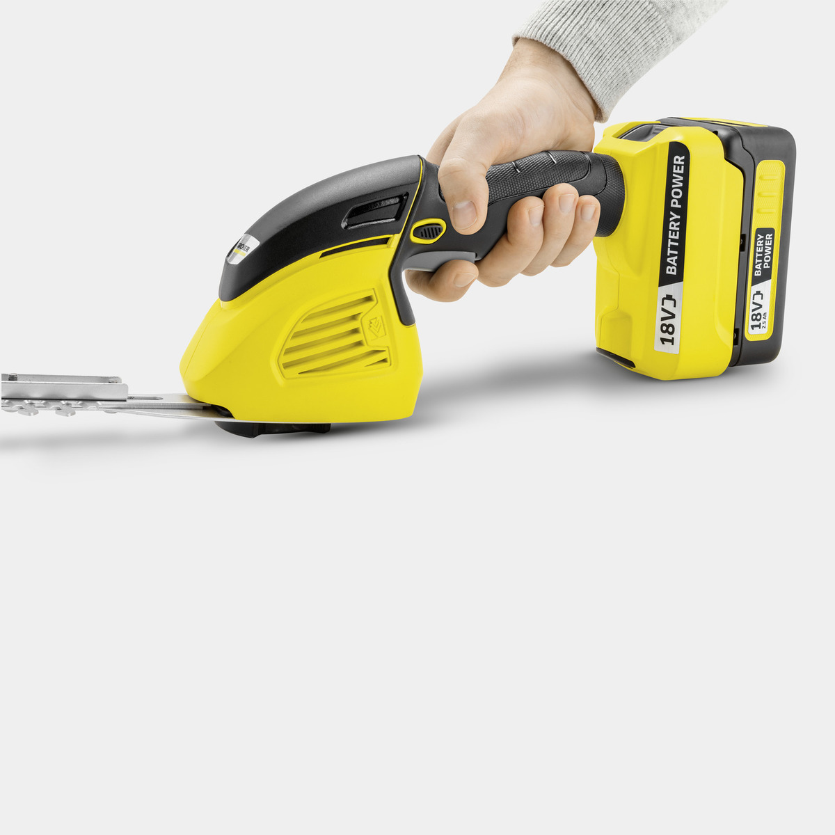 La mano de una persona sosteniendo un cortasetos amarillo y negro, listo para cortar. La batería es visible.
