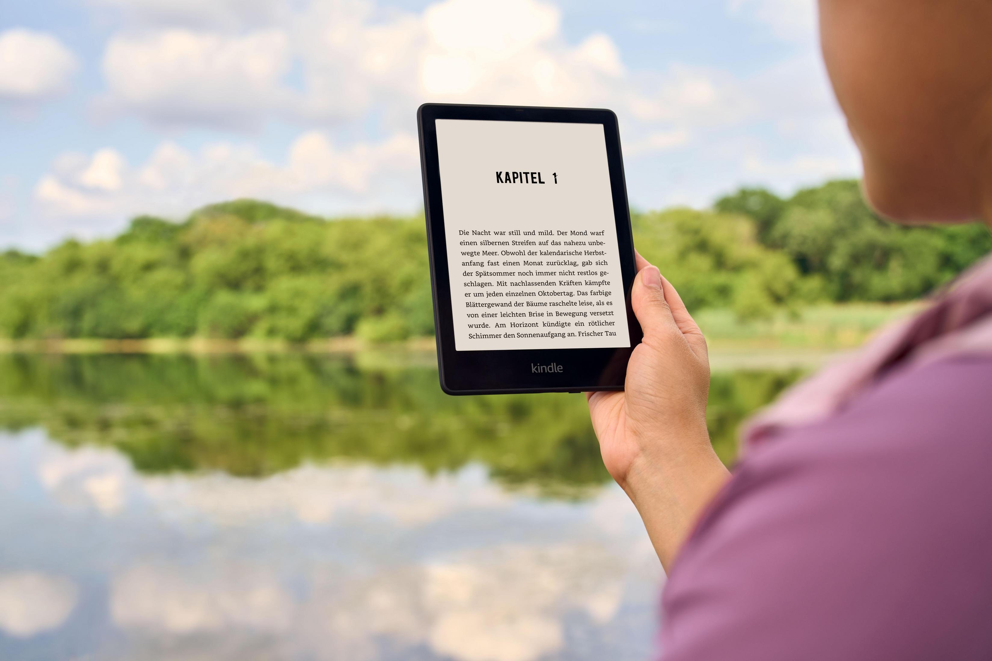 Person hält Kindle-Lesegerät. Der Bildschirm zeigt Text. Hintergrund ist See und Bäume. Person trägt pinkes Hemd.