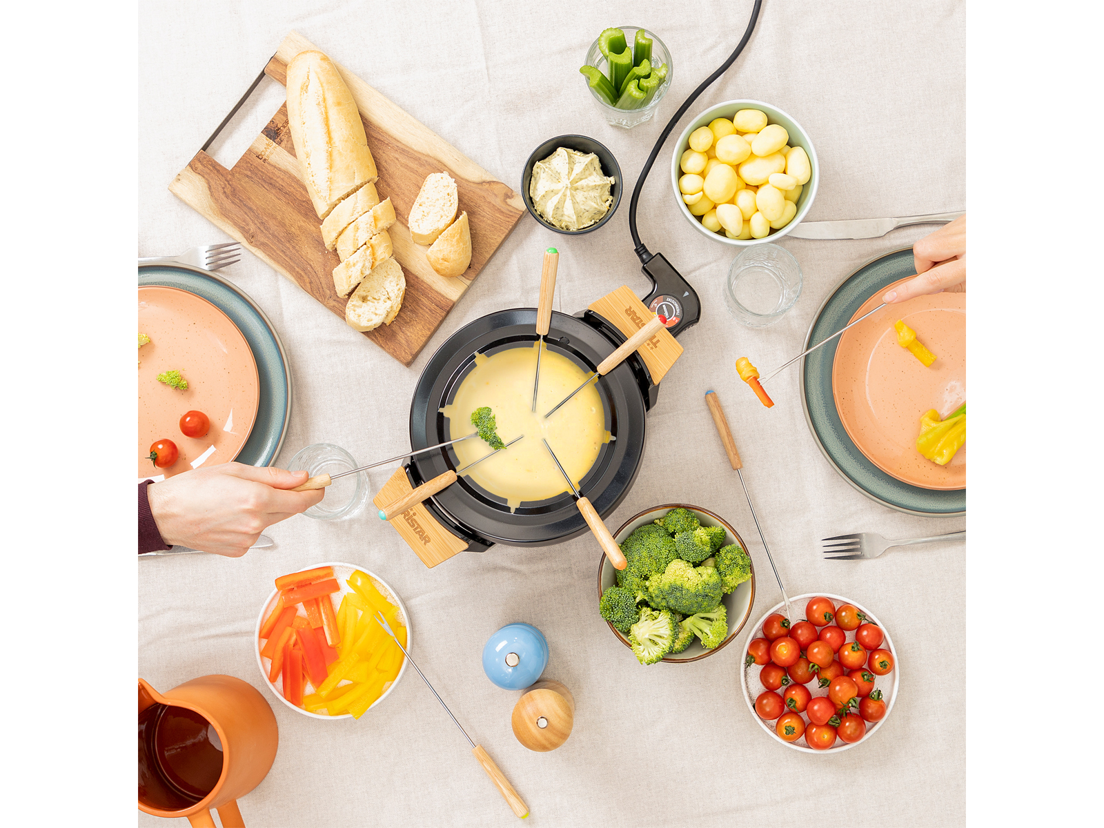 Un juego de fondue en una mesa con alimentos alrededor, gente alcanzando. La marca Tristar es visible.